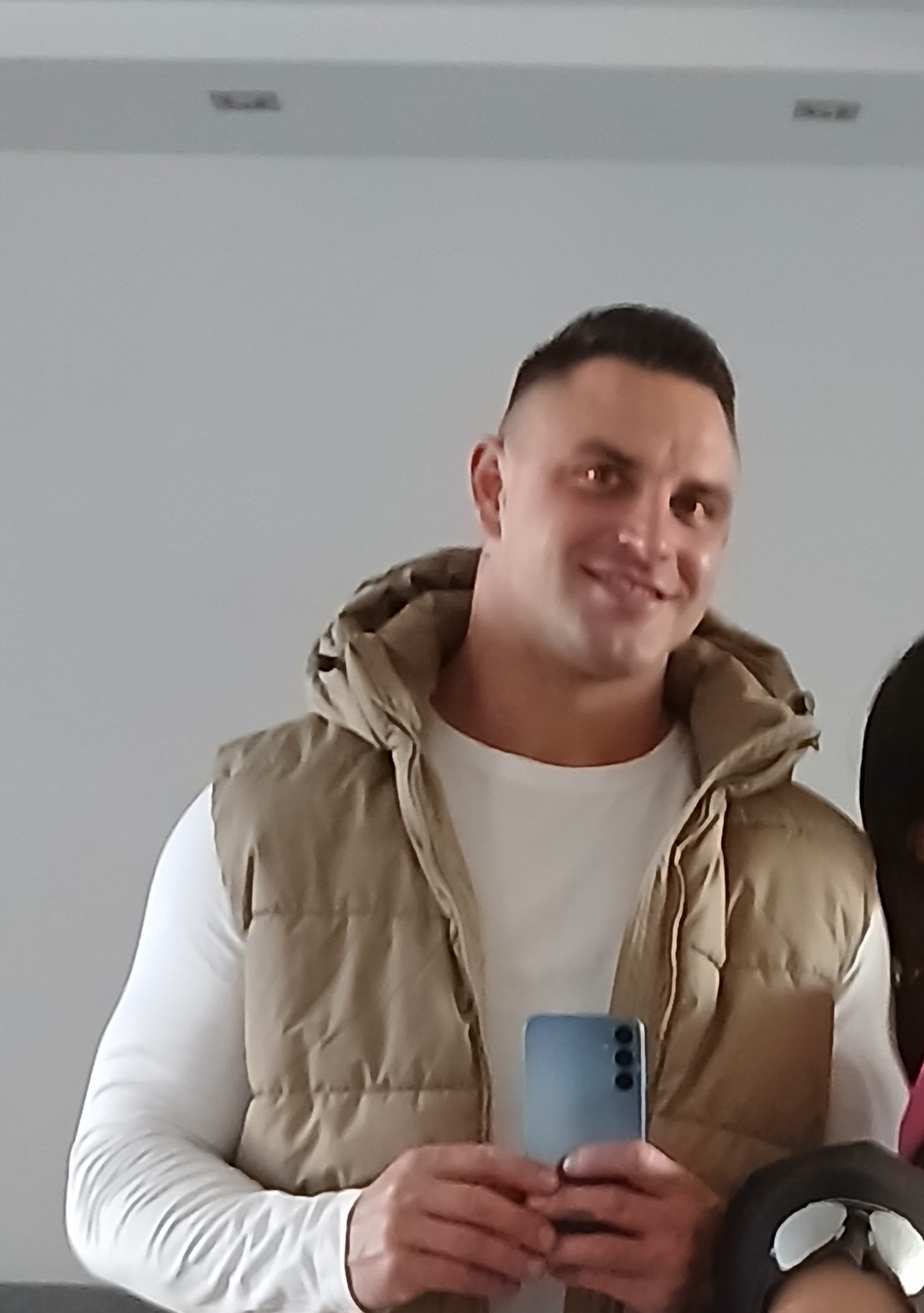 Uśmiechnięty mężczyzna w beżowej kamizelce puchowej z kapturem i białej koszulce z długim rękawem trzymający smartfon na tle jasnej ściany.