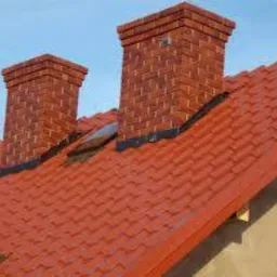 Czerwony dach z dachówki ceramicznej z dwoma kominami z cegły klinkierowej oraz oknem dachowym, widoczne detale obróbek blacharskich.