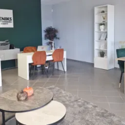 Jasne biuro nieruchomości z logo 'Feniks', biurkami, krzesłami i dekoracjami. Nowoczesne wnętrze, stół z zielonymi krzesłami, dywan i stoliki kawowe.