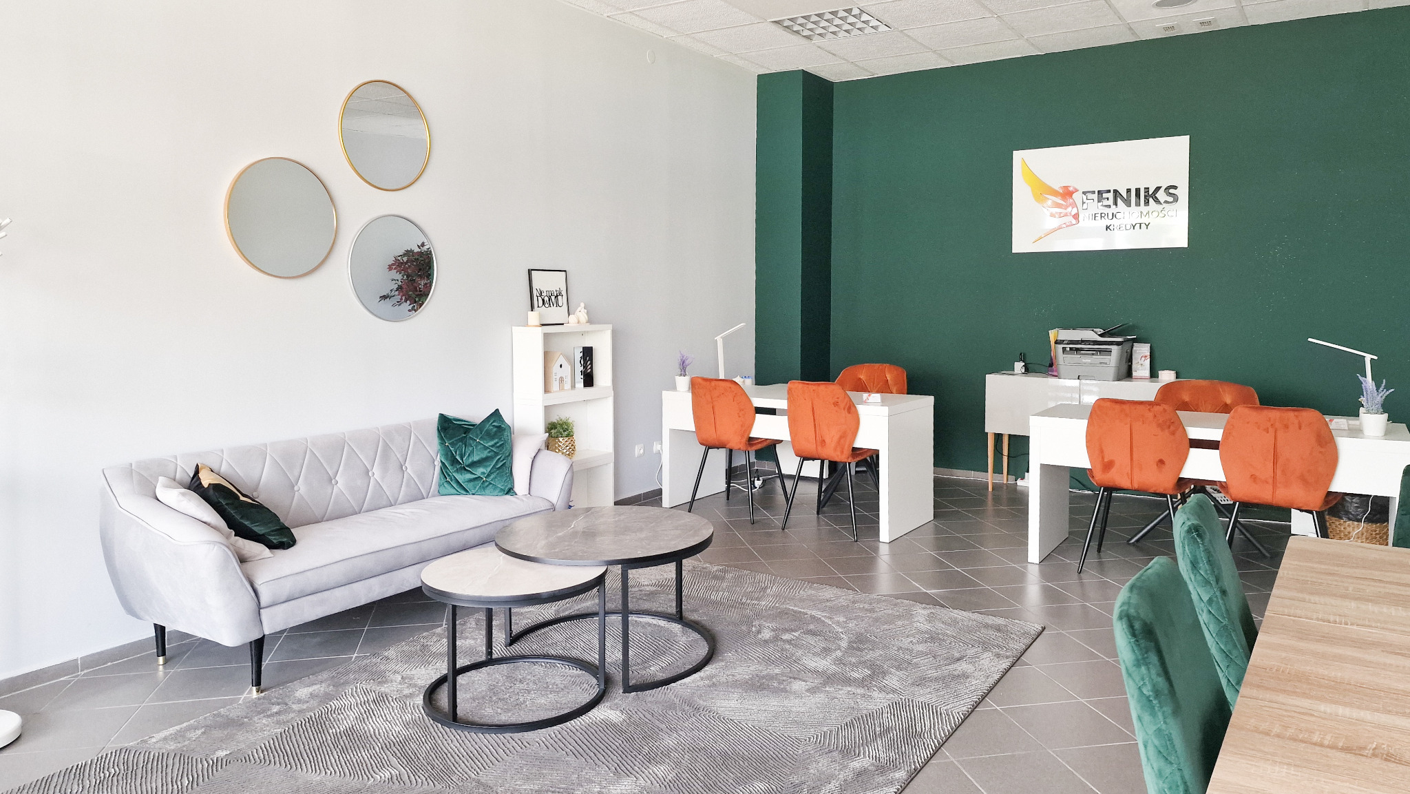 Wnętrze biura nieruchomości z sofą, stolikami kawowymi i biurkami z krzesłami. Na ścianie logo firmy Feniks Nieruchomości i Kredyty. Nowoczesny design, stonowana kolorystyka.