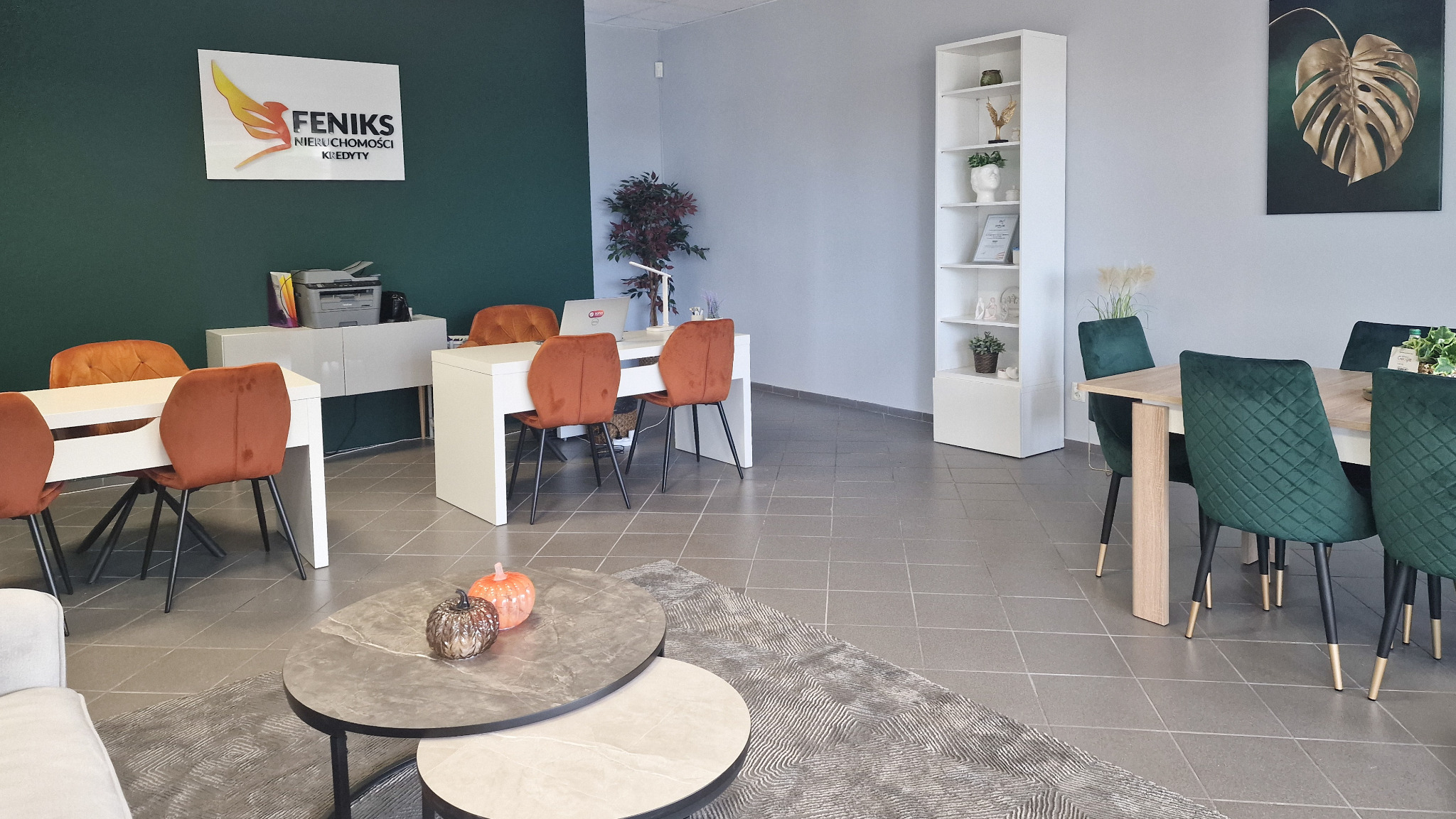 Jasne biuro nieruchomości z logo 'Feniks', biurkami, krzesłami i dekoracjami. Nowoczesne wnętrze, stół z zielonymi krzesłami, dywan i stoliki kawowe.