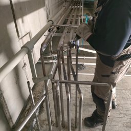 BR-SPAW Usługi spawalnicze mobilne - Pracownik w kombinezonie łączy elementy stalowej konstrukcji rurociągu za pomocą wiertarki, w surowym wnętrzu hali przemysłowej.