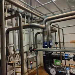 Złożony system rur i zawor&oacute;w w kotłowni, z widocznymi manometrami, pompami i stalową konstrukcją pod sufitem.