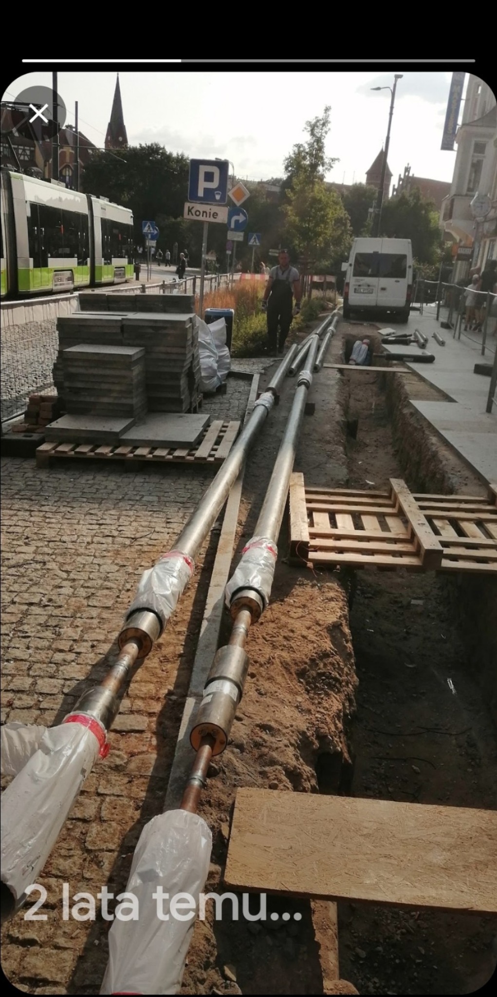 Rury ciepłownicze z kompensatorami ułożone na ziemi obok wykopu w trakcie budowy sieci ciepłowniczej na ulicy miejskiej, w tle tramwaj i budynki.