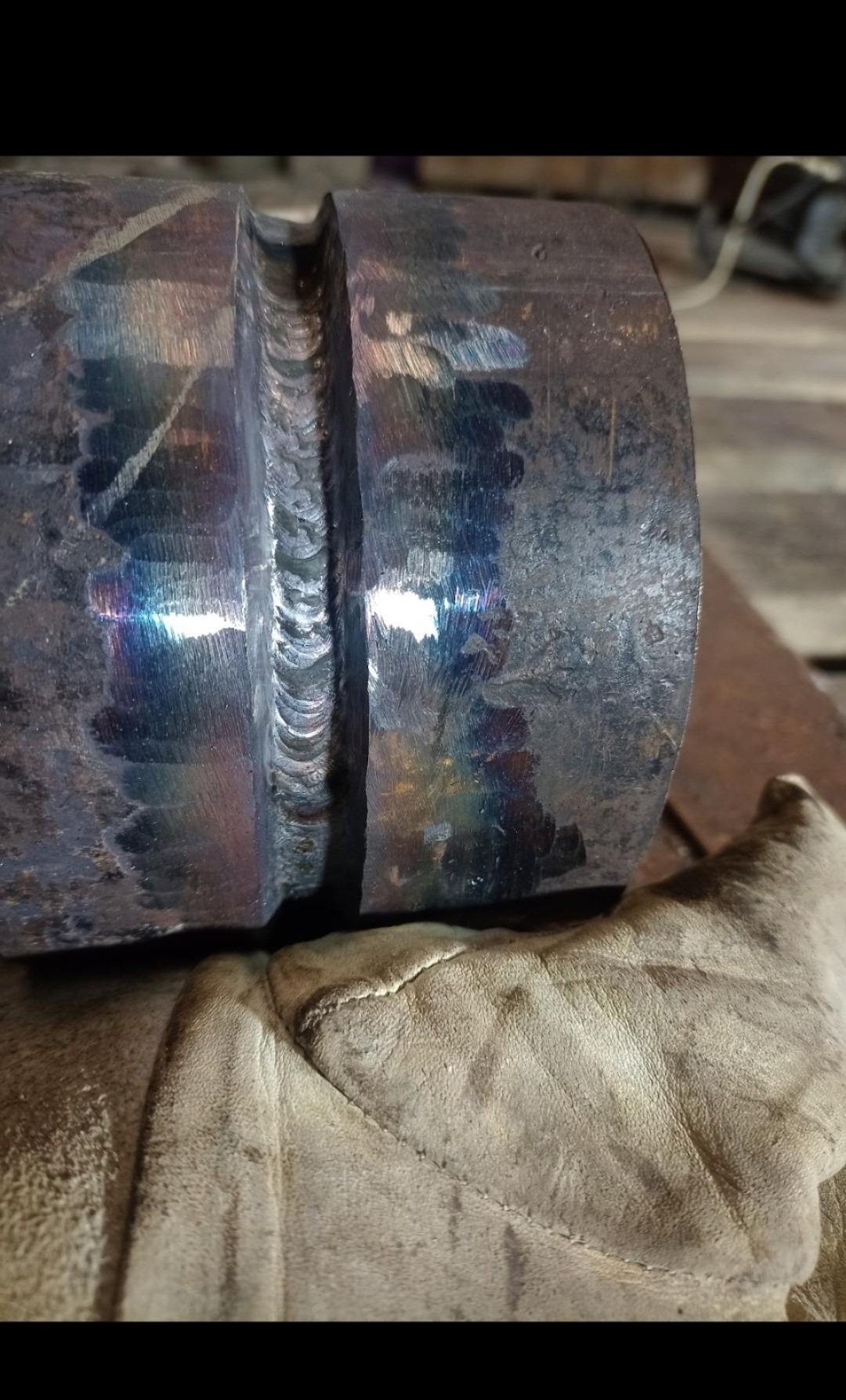 Detaliczne ujęcie spoiny na grubościennej rurze stalowej, widoczne tęczowe przebarwienia termiczne i rękawica ochronna.