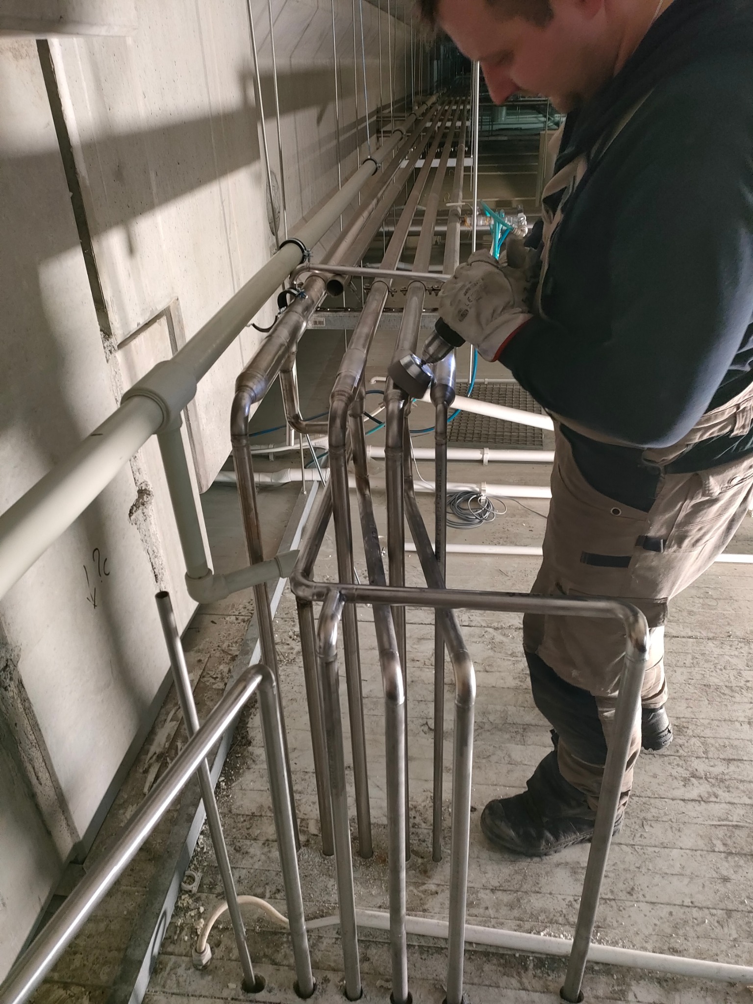 Pracownik w kombinezonie łączy elementy stalowej konstrukcji rurociągu za pomocą wiertarki, w surowym wnętrzu hali przemysłowej.