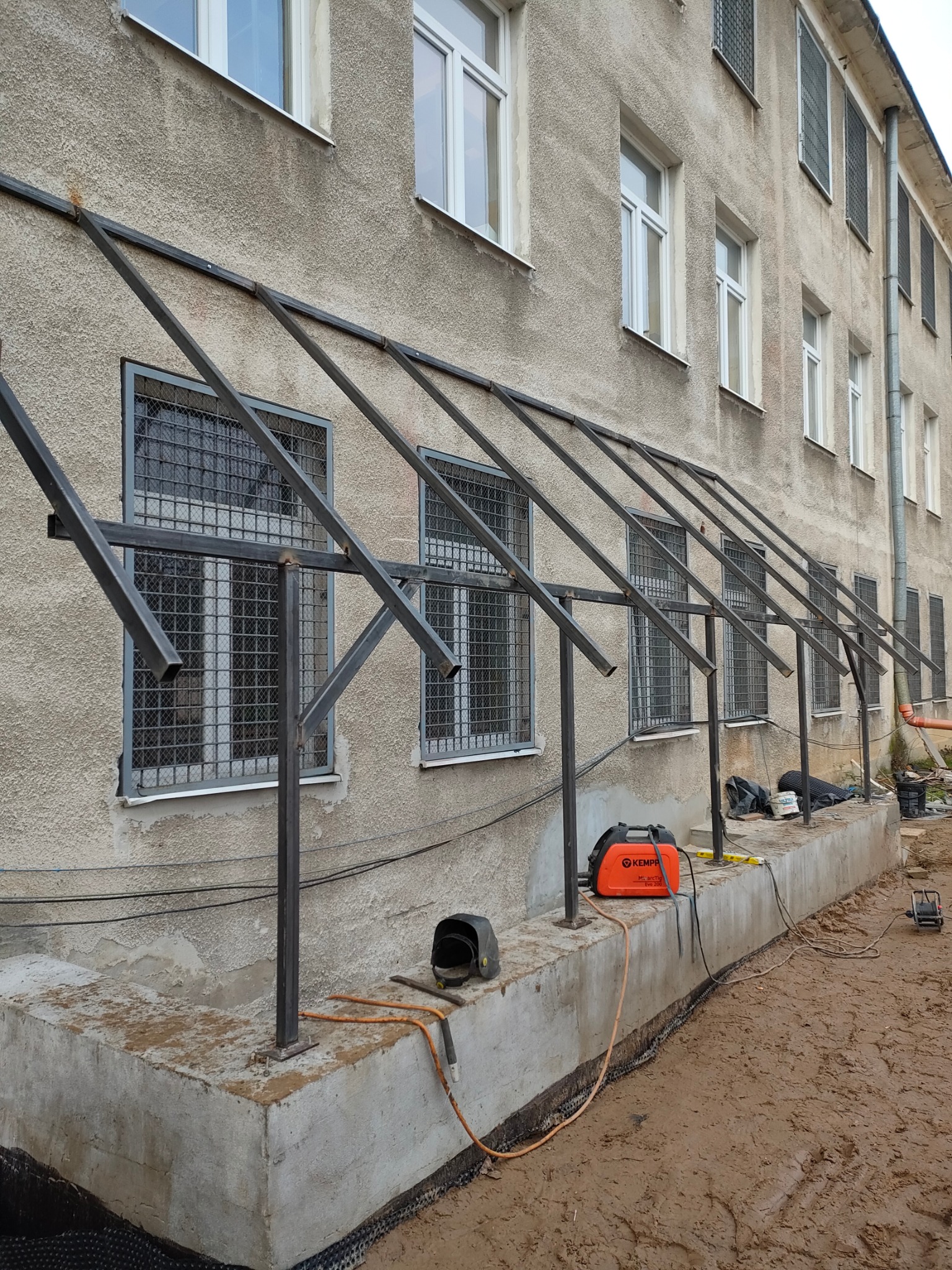 Montaż stalowej konstrukcji zadaszenia nad oknami budynku, widoczny spawacz, spawarka Kemppi, przewody, elementy metalowe oraz podłoże z ziemią.
