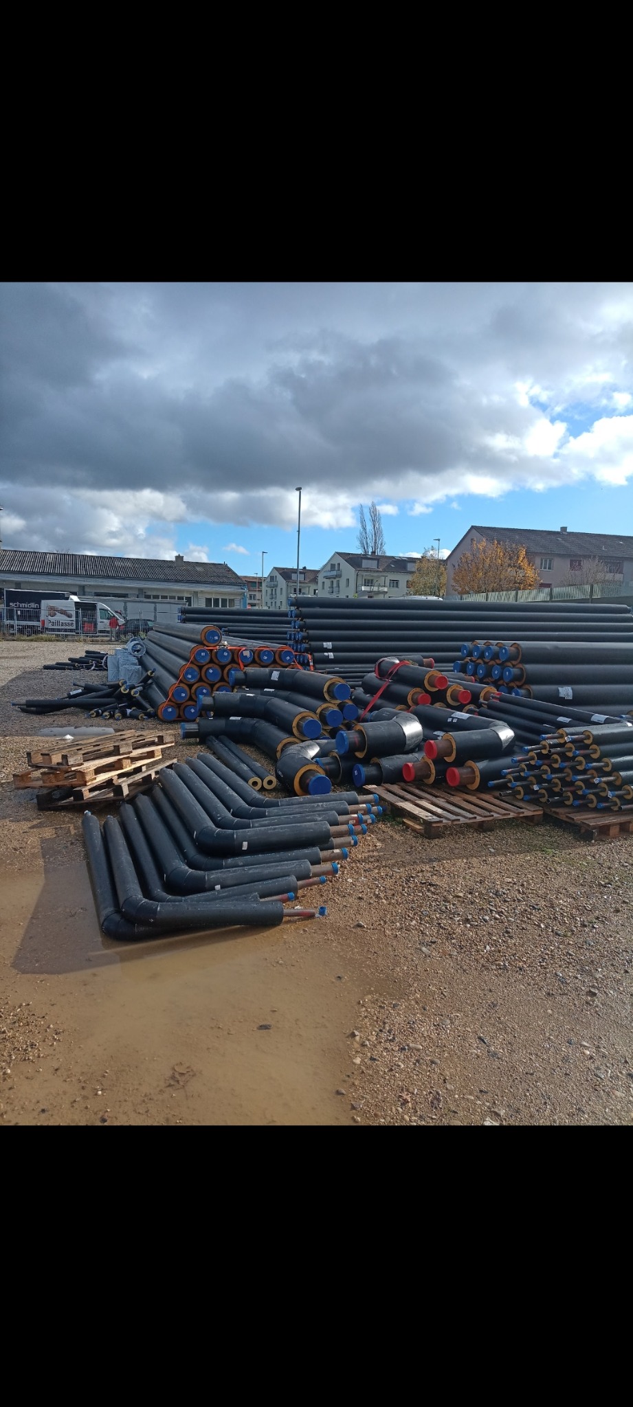 Skladowane rury preizolowane z niebieskimi i czerwonymi zaślepkami na końcach, leżące na paletach i ziemi na placu budowy.