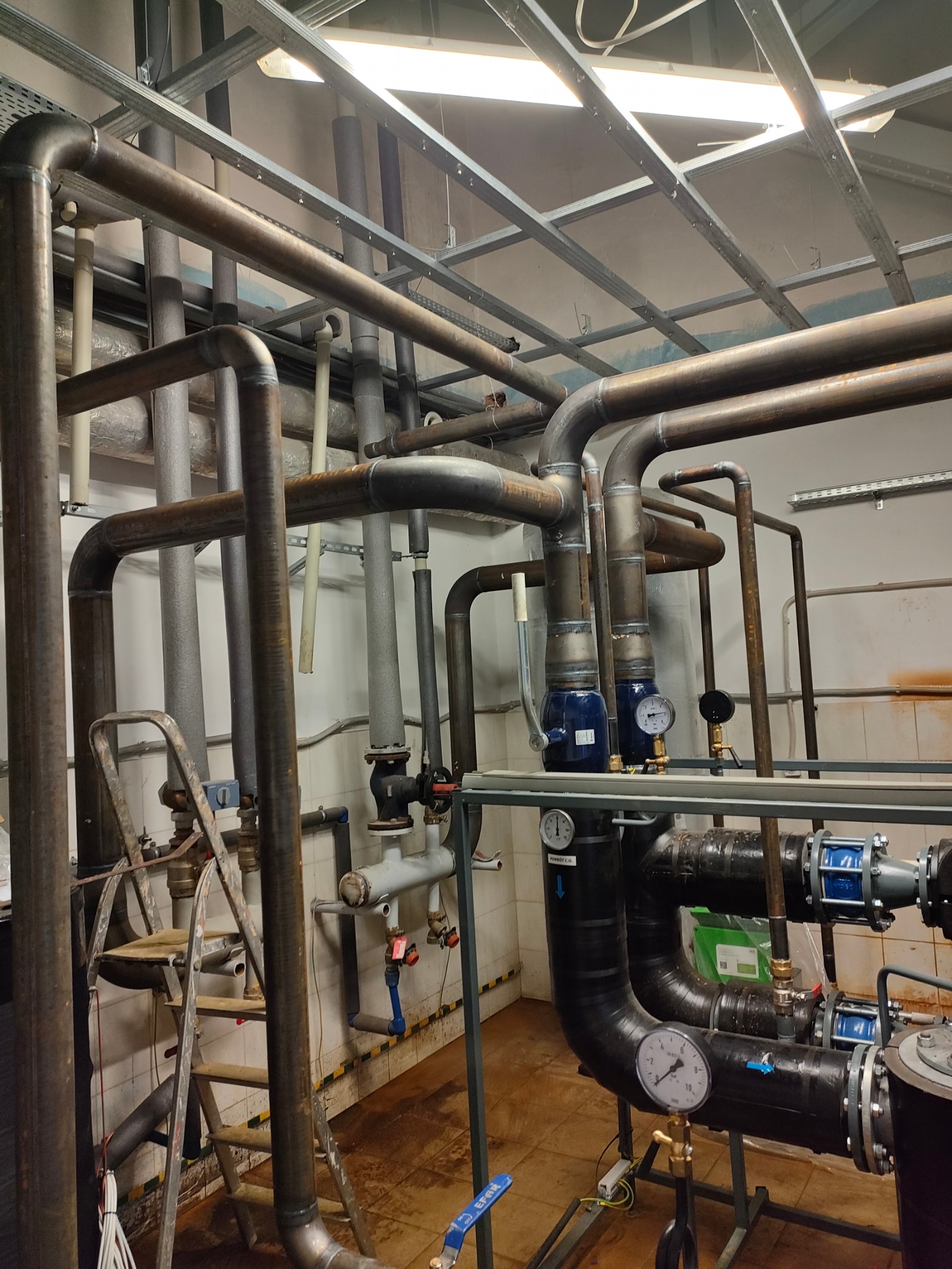 Złożony system rur i zaworów w kotłowni, z widocznymi manometrami, pompami i stalową konstrukcją pod sufitem.