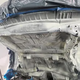 Podwozie samochodu po nałożeniu białej powłoki ochronnej, widoczne elementy konstrukcyjne i zabezpieczenia z folii, ujęcie od dołu.