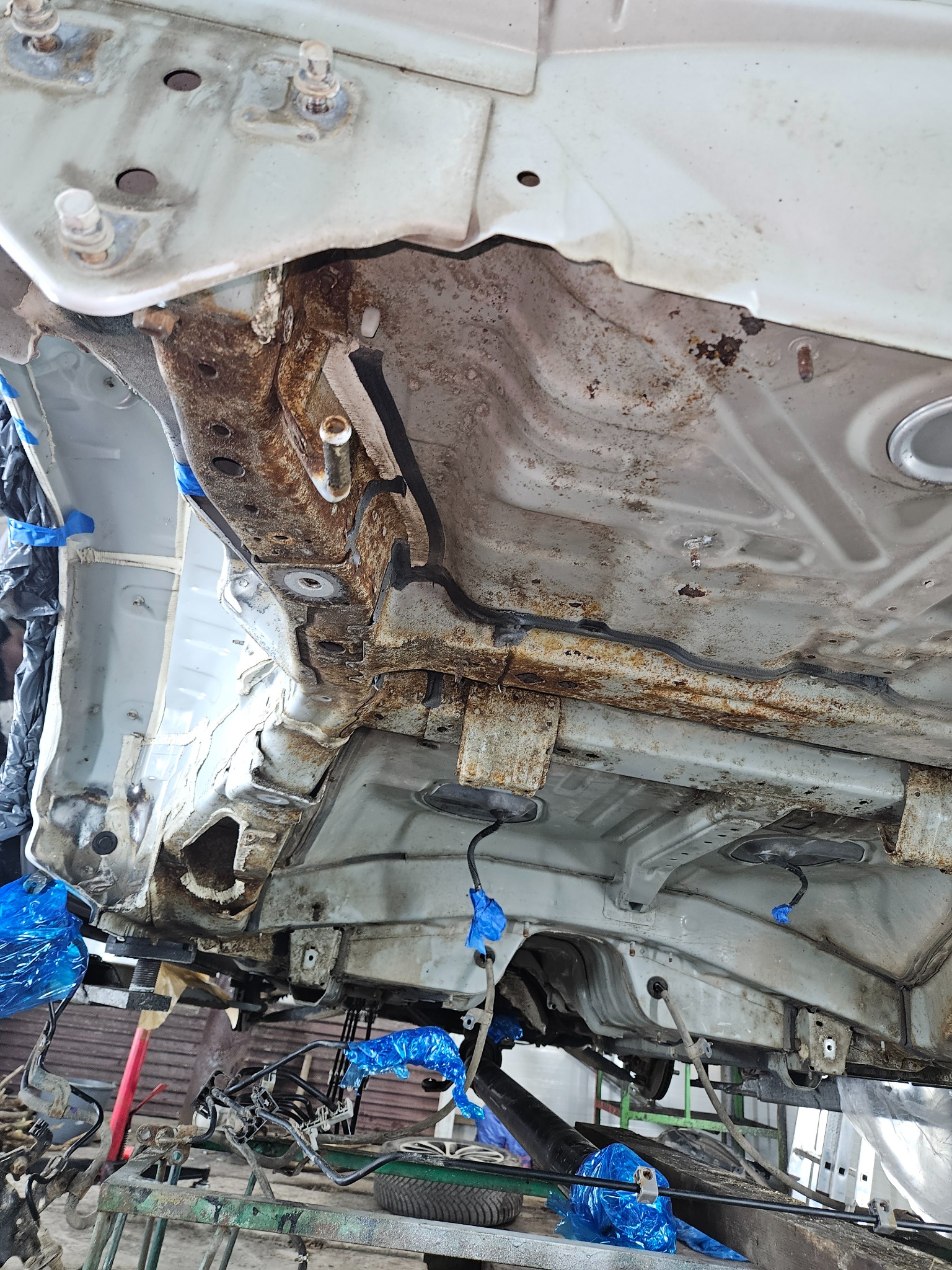 Widok od dołu na podwozie samochodu z widocznymi śladami korozji i elementami zabezpieczonymi folią w kolorze niebieskim, podczas prac warsztatowych.