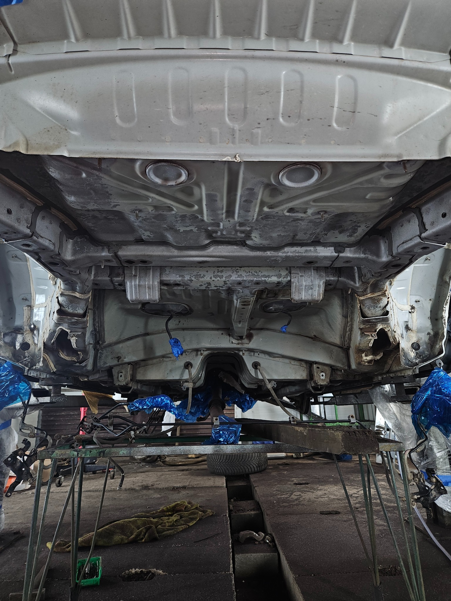 Widok podwozia samochodu na podnośniku warsztatowym, elementy zabezpieczone folią, widoczne ślady korozji i napraw blacharskich.