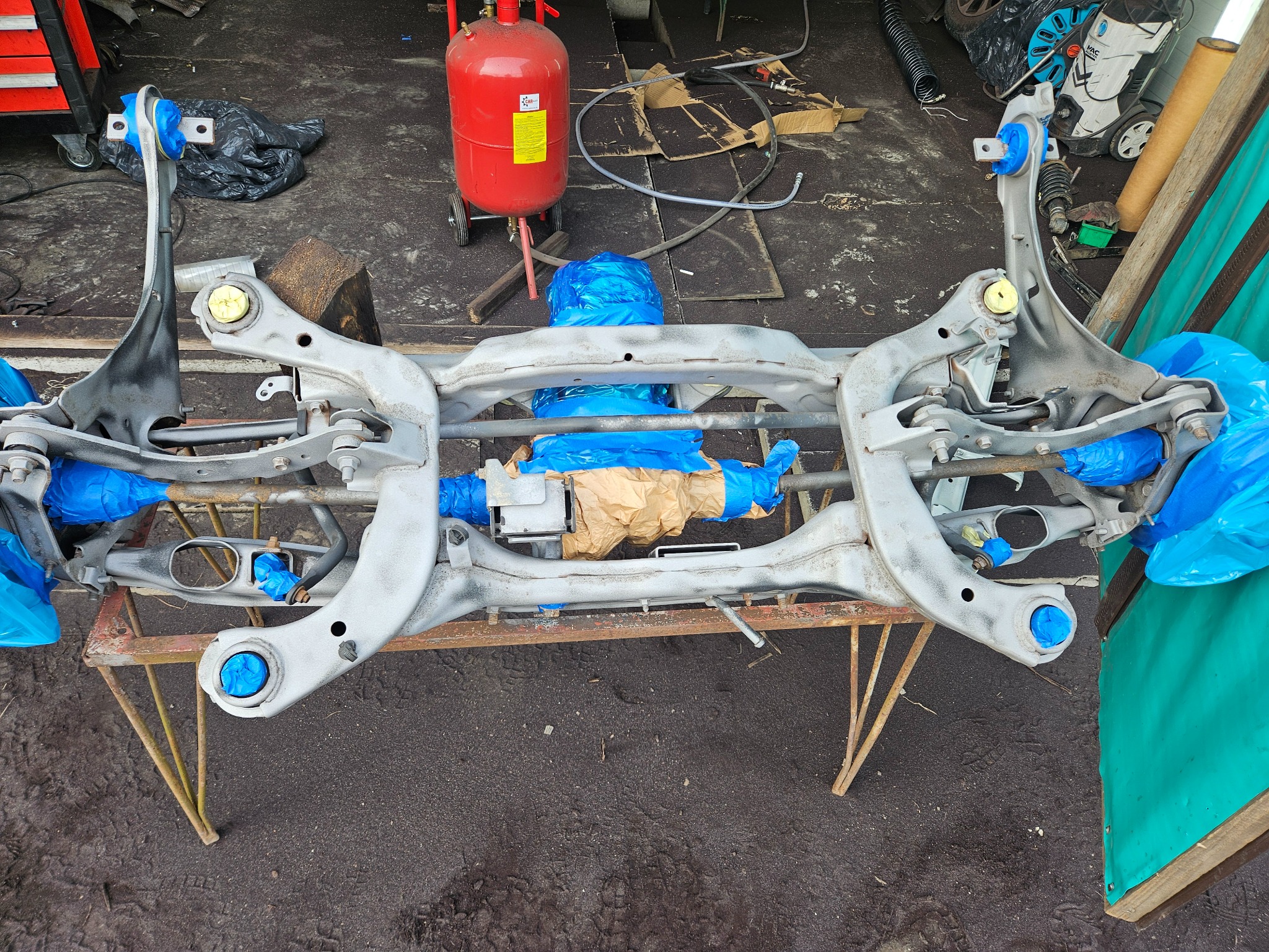 Elementy zawieszenia samochodu po piaskowaniu, przygotowane do malowania, z zabezpieczonymi elementami niebieską folią, na tle warsztatu z piaskarką.