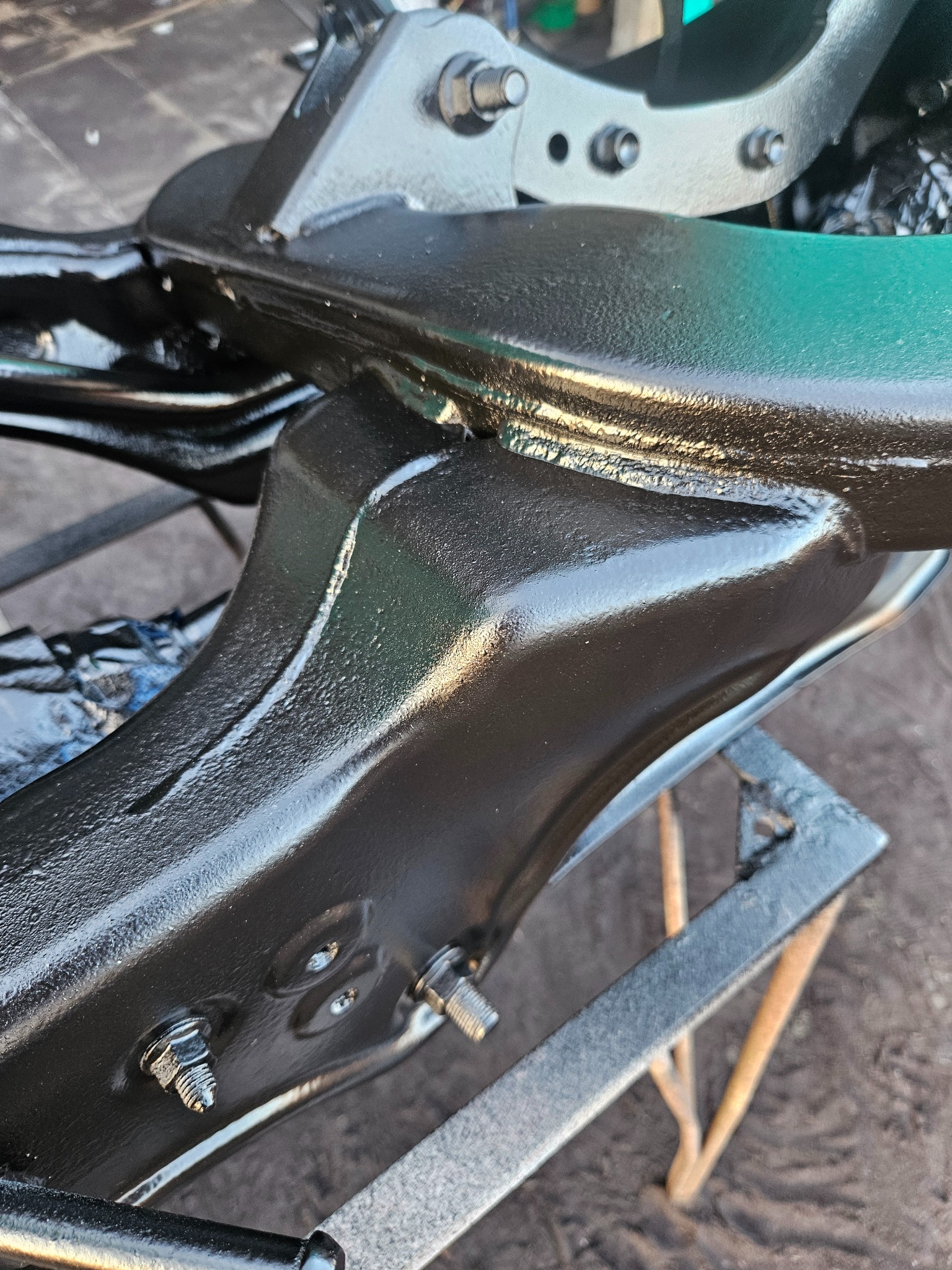 Element podwozia samochodowego po zabezpieczeniu antykorozyjnym, z widocznymi śrubami i mocowaniami, ustawiony na metalowym stojaku.