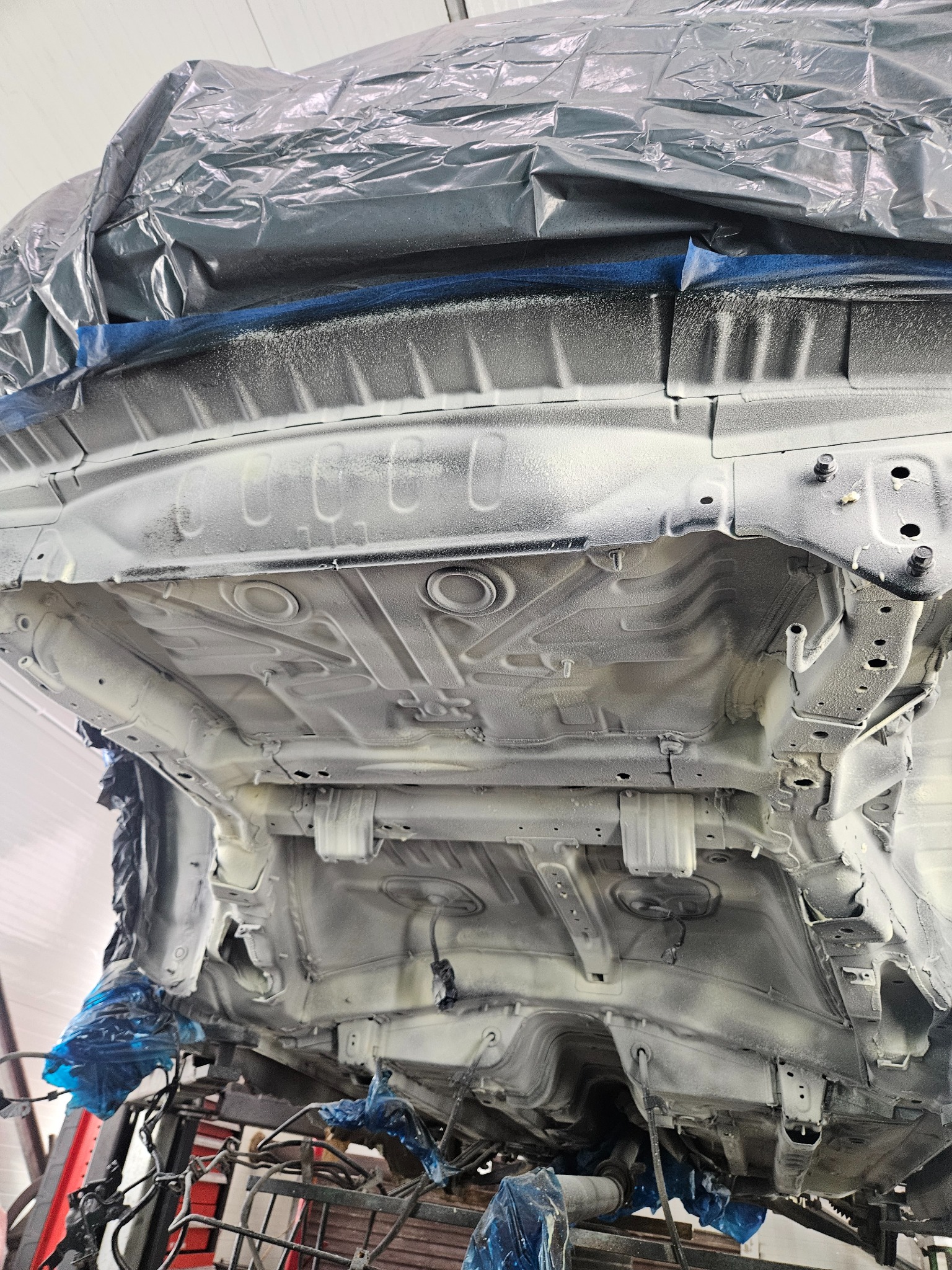 Podwozie samochodu po nałożeniu białej powłoki ochronnej, widoczne elementy konstrukcyjne i zabezpieczenia z folii, ujęcie od dołu.