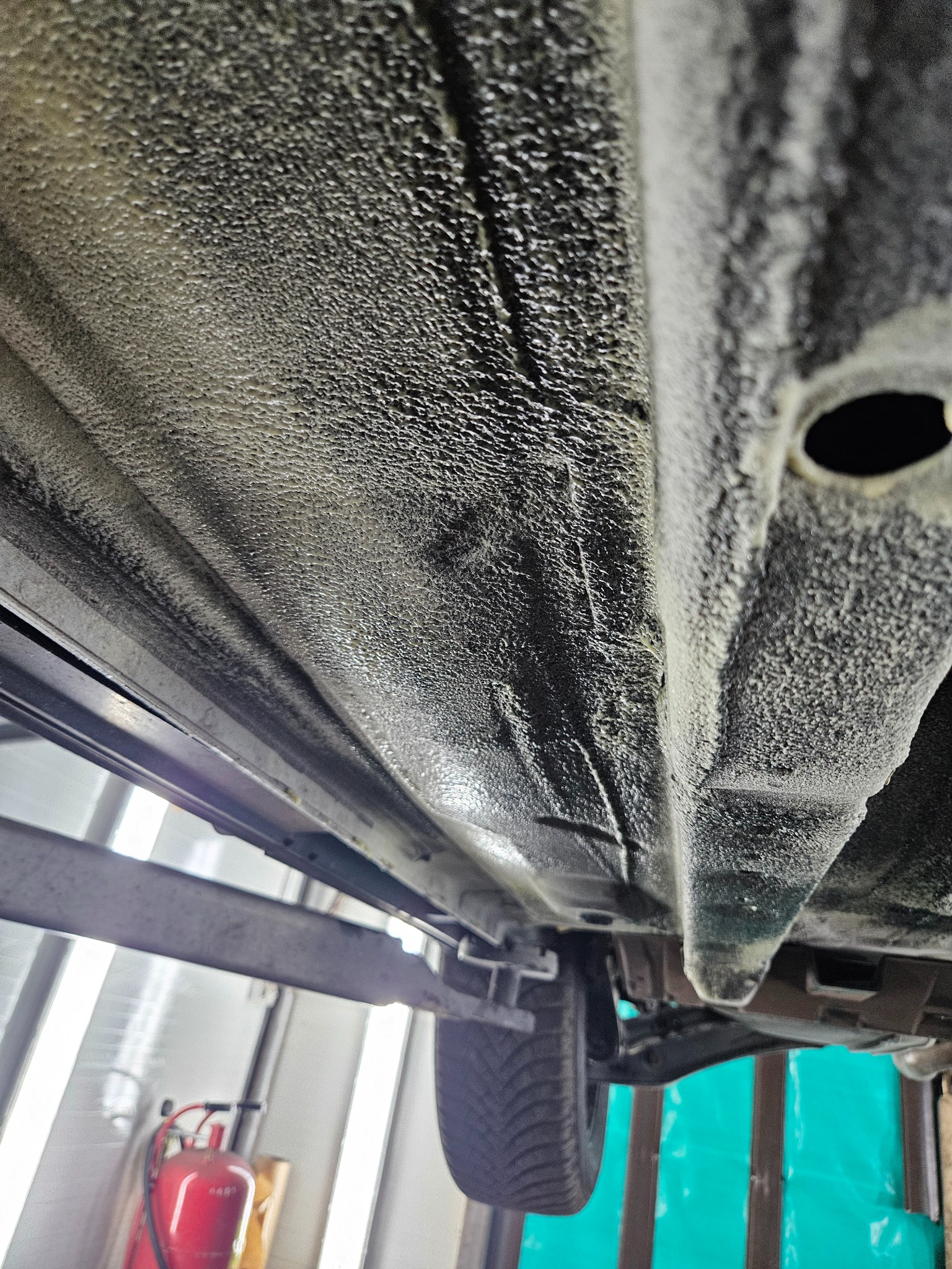 Spód samochodu na podnośniku po nałożeniu grubej warstwy antykorozyjnej, widoczna struktura baranka oraz fragment opony i podnośnika.