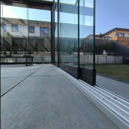 ESTIVO - Szklana konstrukcja z czarnymi profilami i aluminiową szyną dolną, widok z wnętrza na ogród i sąsiedni dom.