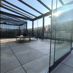 ESTIVO - Szklana zabudowa tarasu z metalową konstrukcją nośną, widoczny system przesuwny szyb, meble ogrodowe i dekoracje na stoliku kawowym.