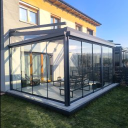 ESTIVO - Szklana zabudowa tarasu z przesuwnymi drzwiami, widoczne meble ogrodowe i kosiarka w środku, na zewnątrz trawnik i fragment domu w słoneczny dzień.