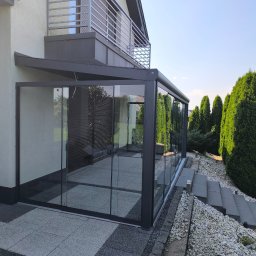 ESTIVO - Nowoczesna, szklana zabudowa tarasu z szarą ramą, przylegająca do budynku z balkonem z metalową balustradą. Widoczne schody i ozdobne tuje w ogrodzie.