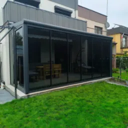 Szklana zabudowa tarasu z ciemnymi ramami, widok z zewnątrz na zielony trawnik i zabudowę domu z balkonem.