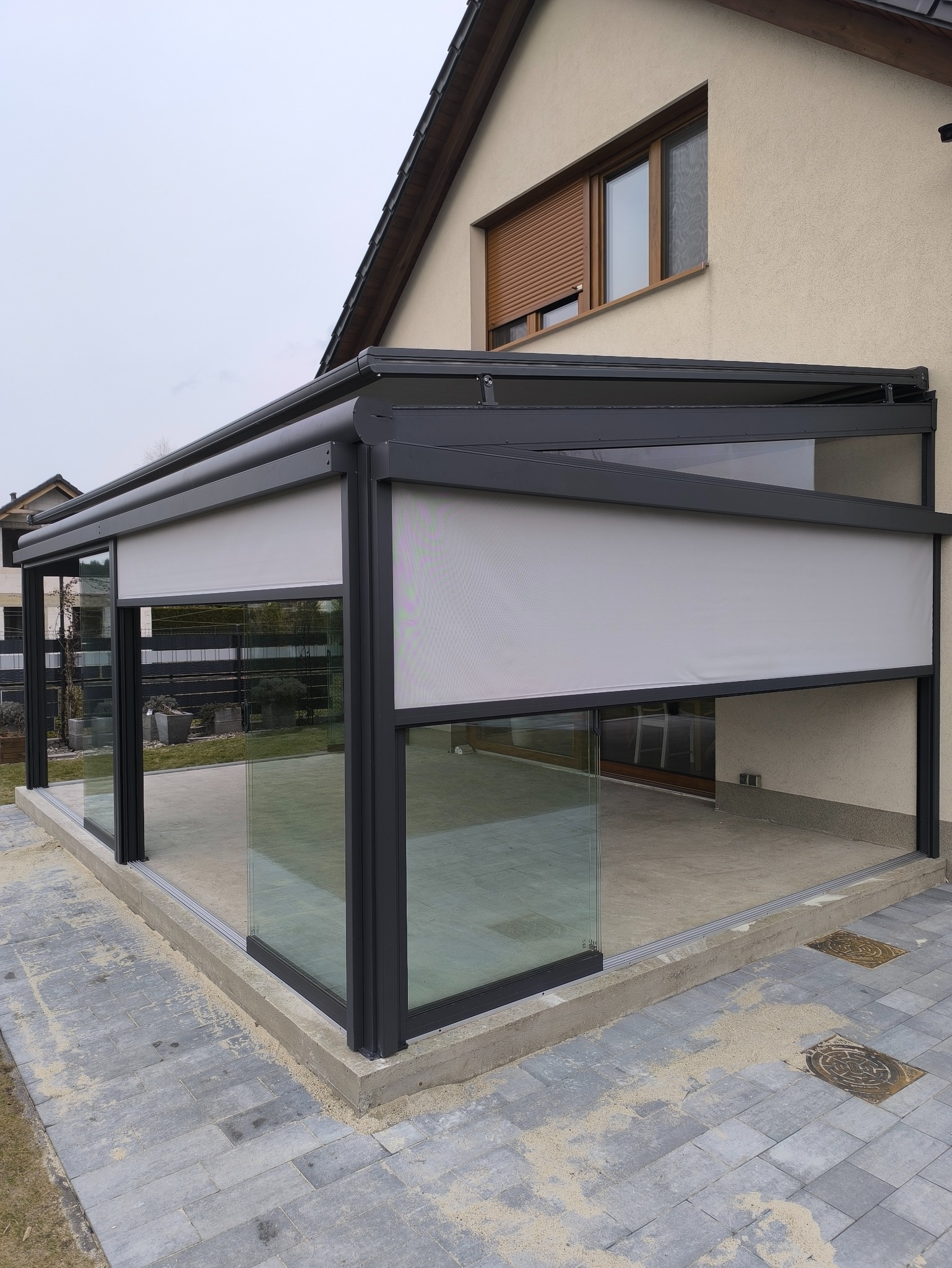 Nowoczesna pergola metalowa z szklanymi ścianami i roletami przeciwsłonecznymi w Bytomiu, przylegająca do budynku z brązowymi roletami okiennymi.