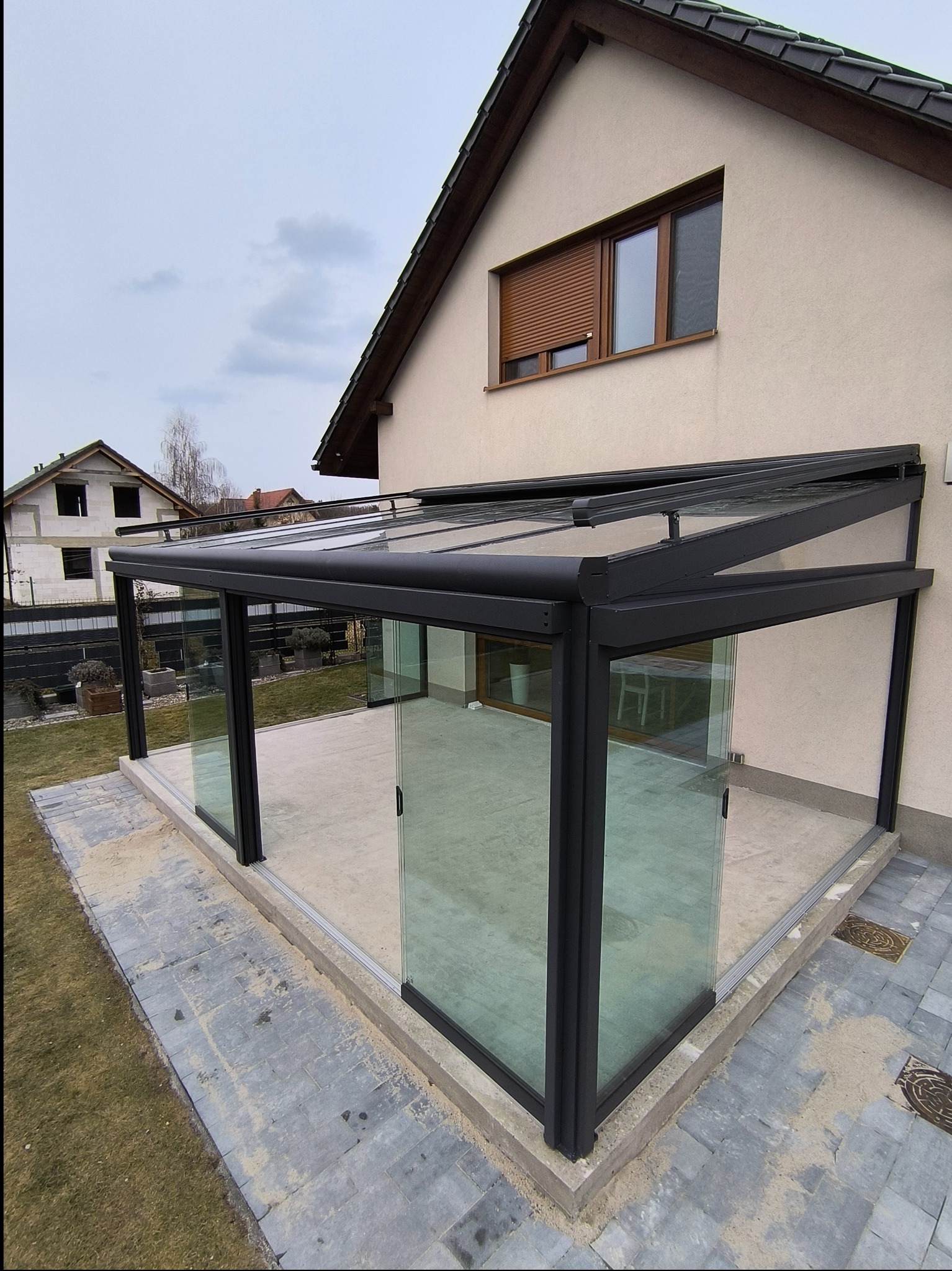 Nowoczesna, przeszklona konstrukcja z czarną ramą, prawdopodobnie weranda lub zabudowany taras, przylegająca do budynku z brązowymi roletami zewnętrznymi.