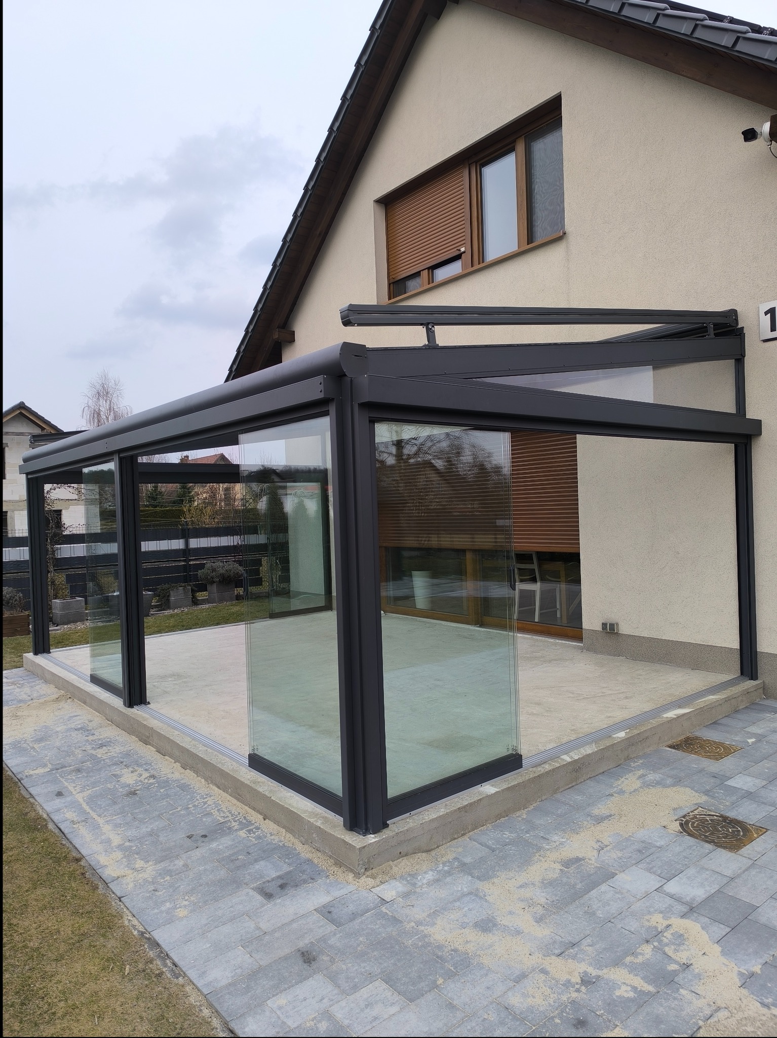 Nowoczesna, przeszklona konstrukcja z czarną ramą, przylegająca do domu z beżową elewacją i brązowymi roletami, otoczona kostką brukową.
