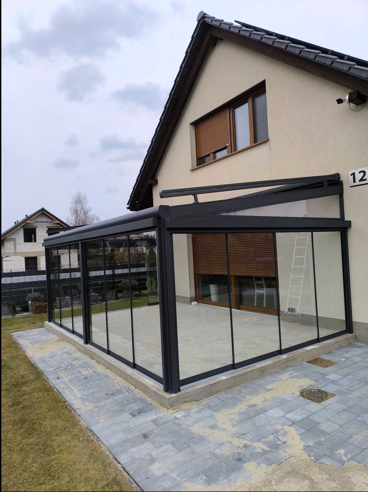 Nowoczesna, przeszklona konstrukcja z czarnymi ramami, przylegająca do ściany domu z numerem 12, z widoczną drabiną od wewnątrz i kostką brukową wokół.
