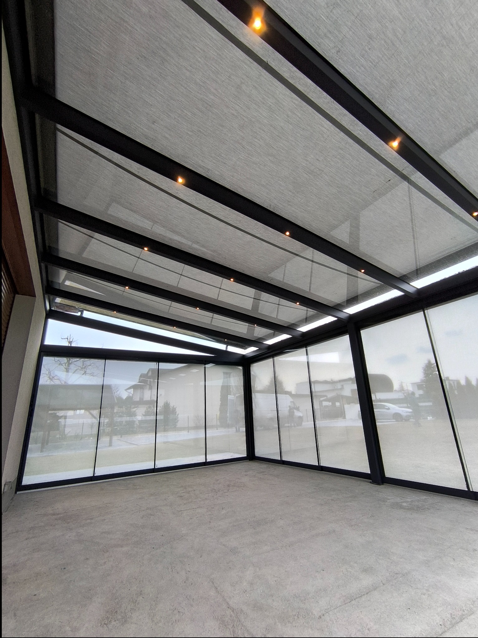 Zadaszona przestrzeń z metalową konstrukcją, szklanymi panelami i roletami, oświetlona punktowymi lampami, widok z dołu na sufit.