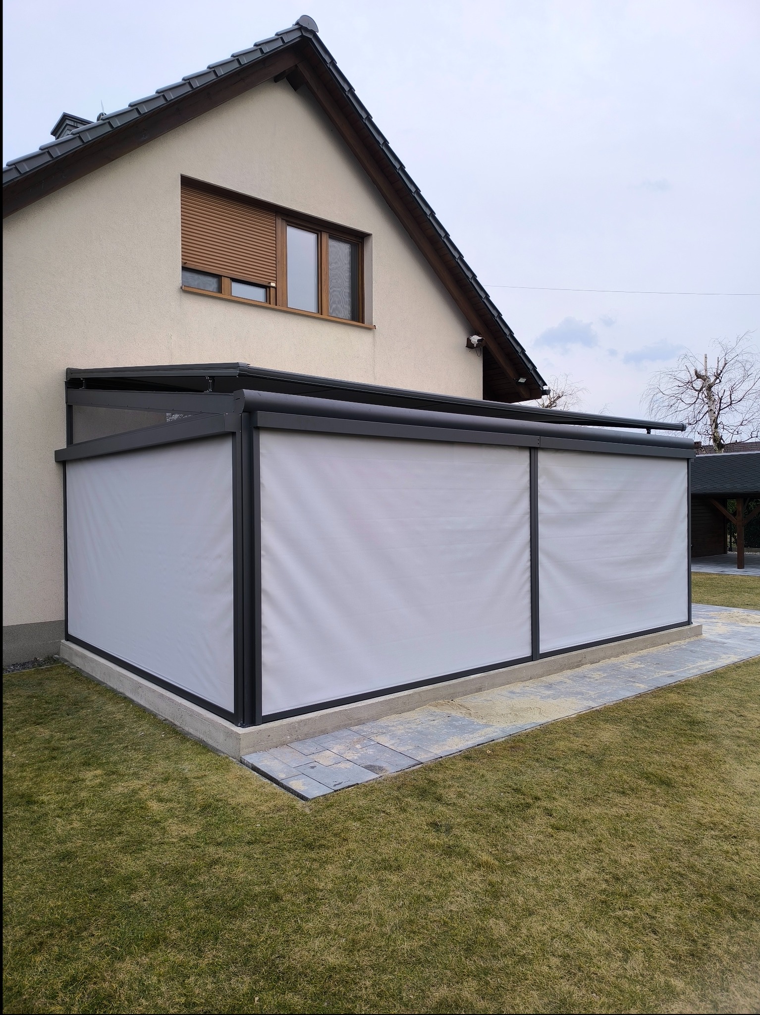 Metalowa pergola przy domu z zasłoniętymi ścianami bocznymi, widok z zewnątrz na trawnik i fragment elewacji domu z oknem.