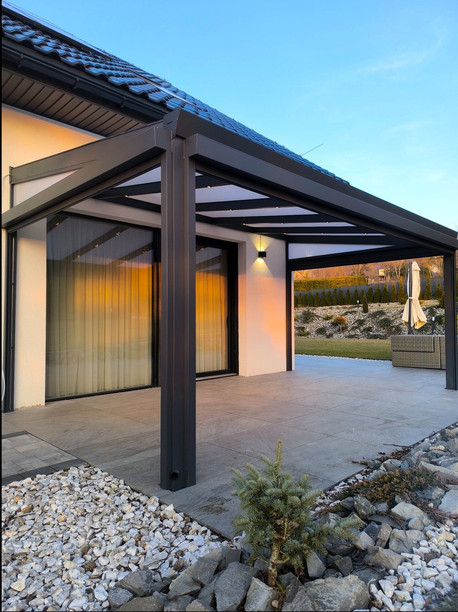 Nowoczesna pergola z metalową konstrukcją, oświetleniem punktowym i przesuwnymi szklanymi panelami, z widokiem na zadbany ogród i kostkę brukową.