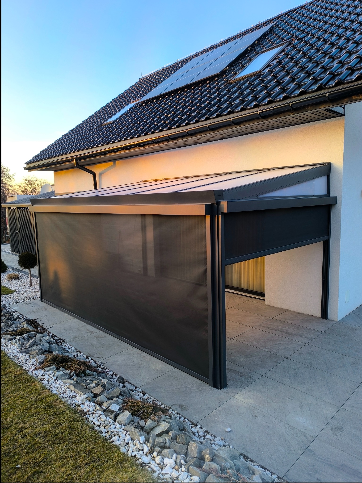 Nowoczesna, grafitowa pergola z częściowo zasłoniętą roletą, przylegająca do białego budynku z panelami słonecznymi na dachu, otoczona minimalistycznym ogrodem z kamieniami.
