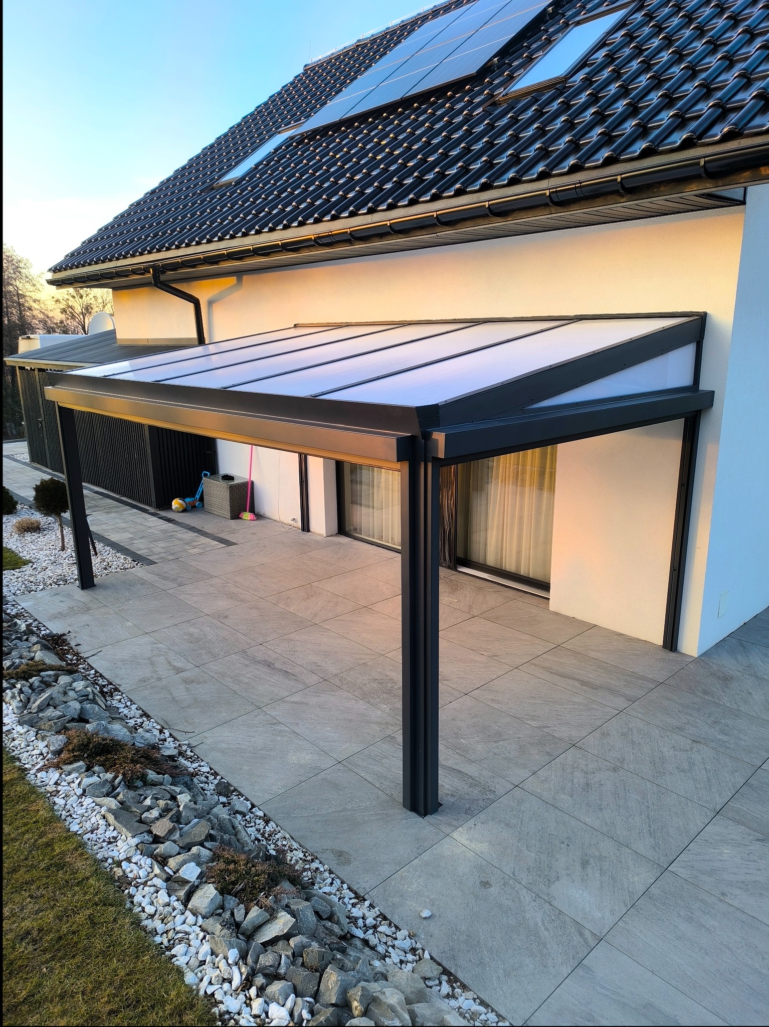 Nowoczesna, czarna pergola z przezroczystym zadaszeniem przylegająca do białej ściany domu z panelami słonecznymi na dachu, otoczona szarą kostką brukową i kamieniami dekoracyjnymi.