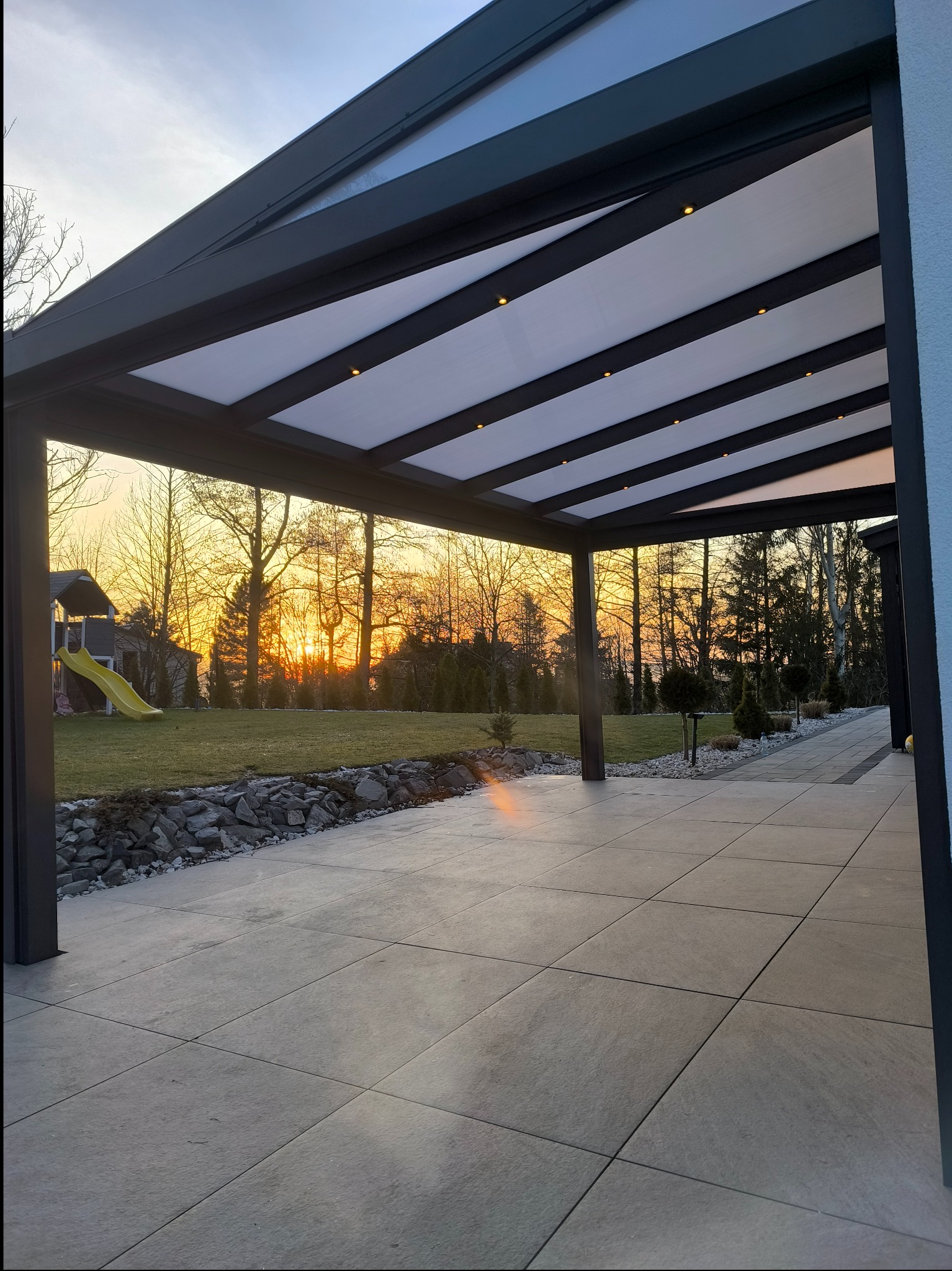 Metalowa pergola z zadaszeniem i oświetleniem LED, widok na ogród z placem zabaw w tle, zachód słońca.