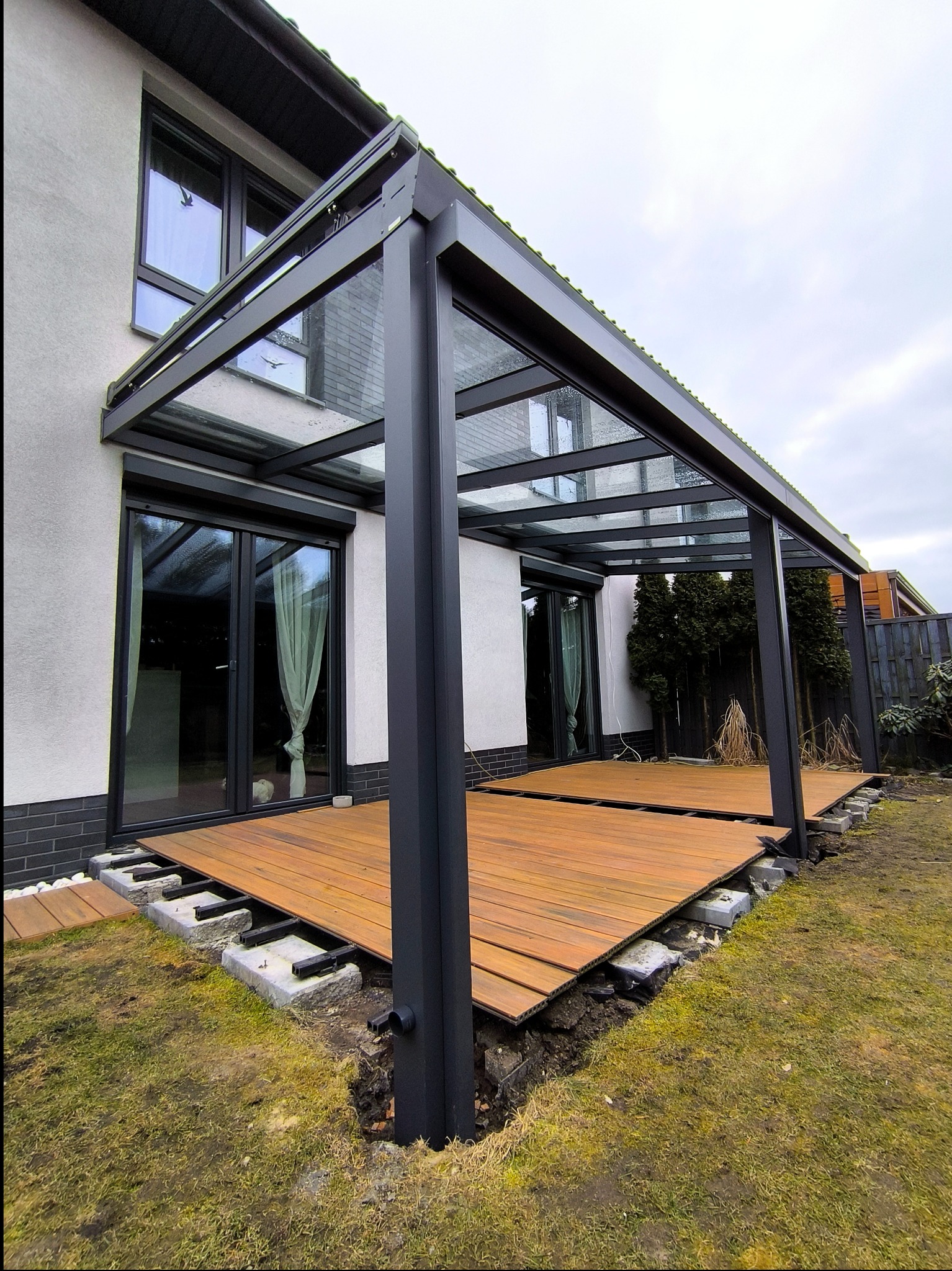 Montaż nowoczesnej, metalowej konstrukcji z przeszklonym dachem, prawdopodobnie pergoli, z widoczną drewnianą podłogą tarasową w trakcie budowy, osadzoną na betonowych bloczkach.