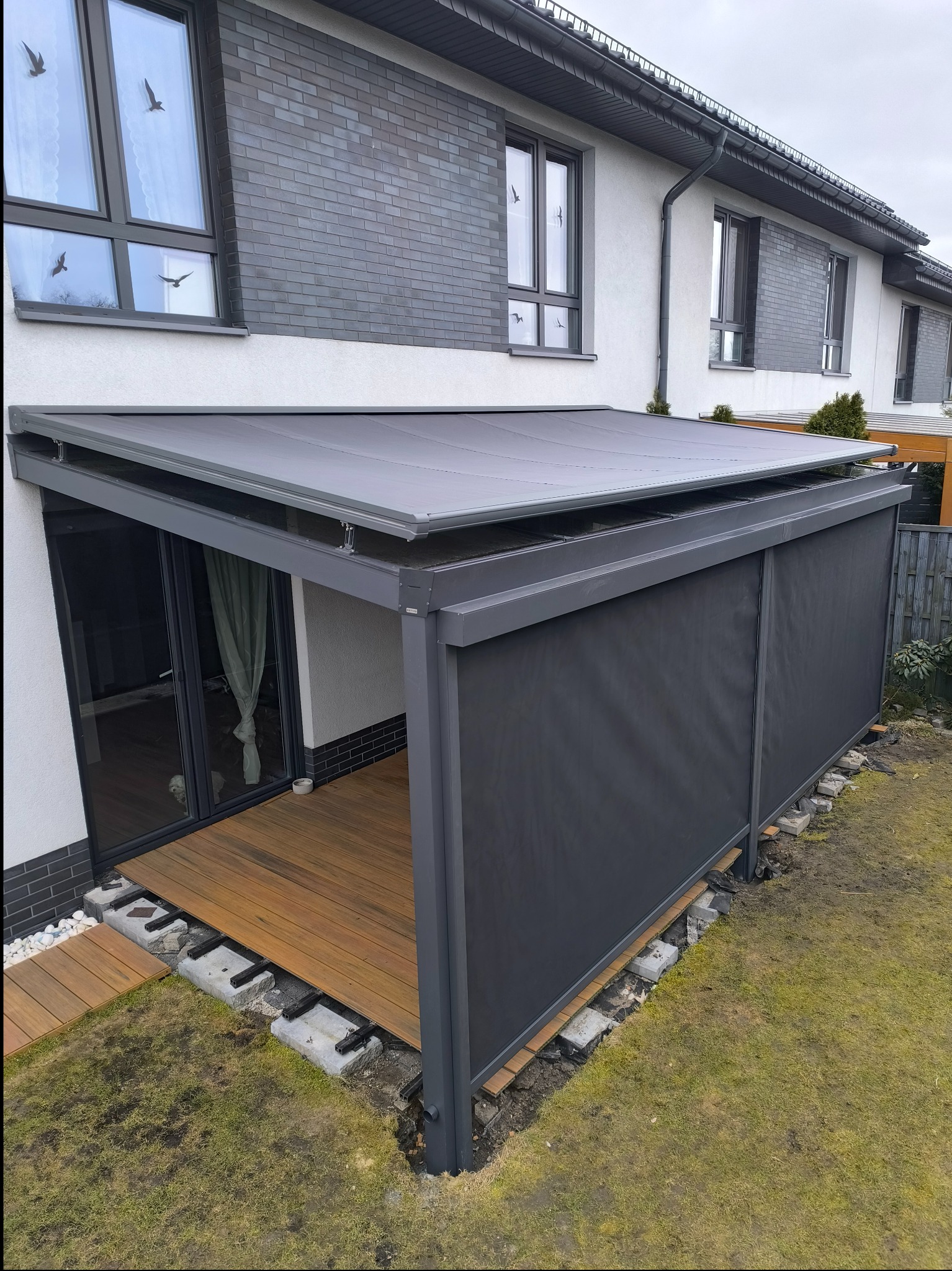 Nowoczesna, szara pergola z rozsuwanymi panelami zacieniającymi, przylegająca do domu z drewnianym tarasem w trakcie budowy.
