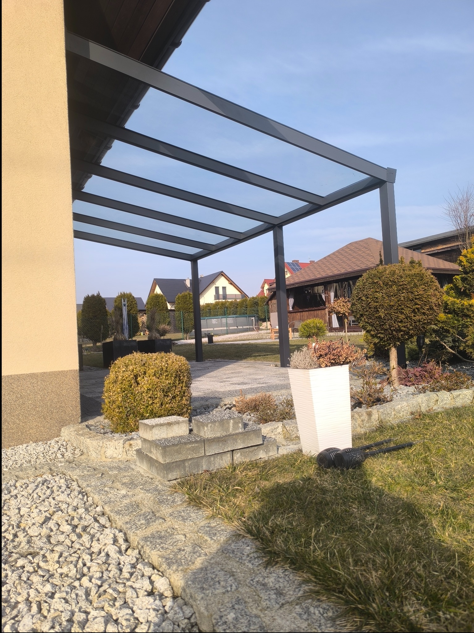 Nowoczesna pergola z metalową konstrukcją i szklanym dachem, widok z zewnątrz na tle zadbanego ogrodu z ozdobnymi krzewami i kostką brukową.