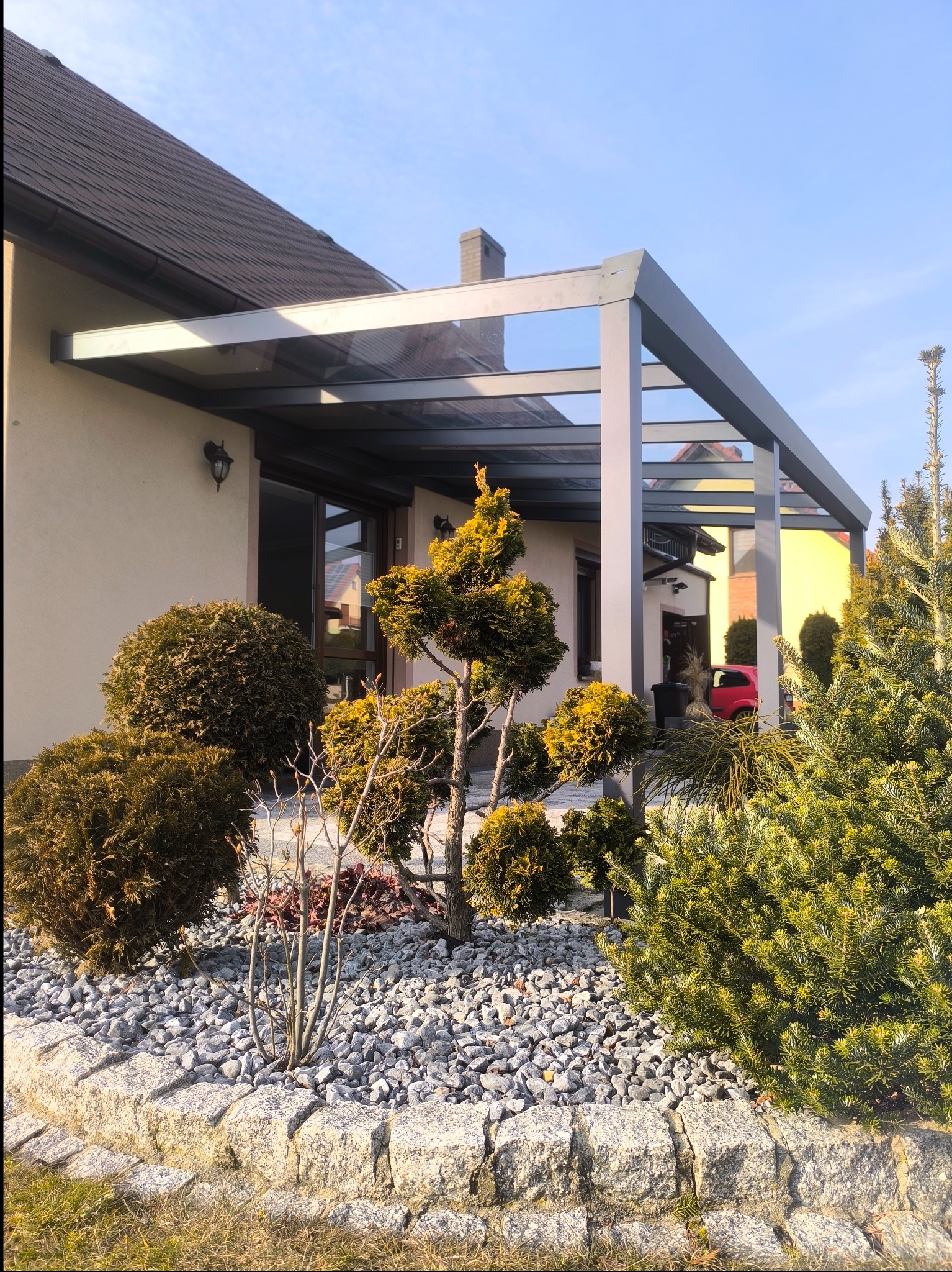 Metalowa konstrukcja z przeszklonym dachem, tworząca zadaszenie tarasu przy domu jednorodzinnym, otoczona ozdobnymi krzewami i kamieniami.