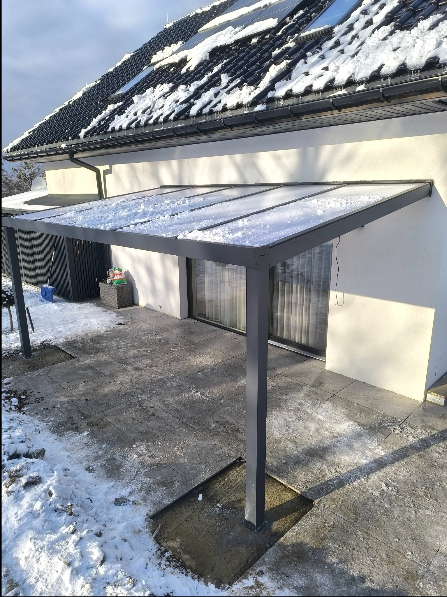 Nowoczesna pergola metalowa przydomowa pokryta śniegiem, z widoczną łopatą do odśnieżania i kostką brukową częściowo zasypaną śniegiem.