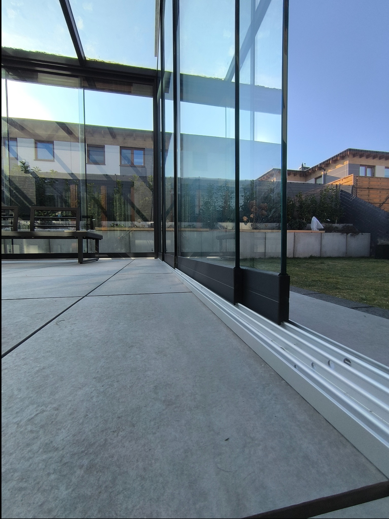 Szklana konstrukcja z czarnymi profilami i aluminiową szyną dolną, widok z wnętrza na ogród i sąsiedni dom.