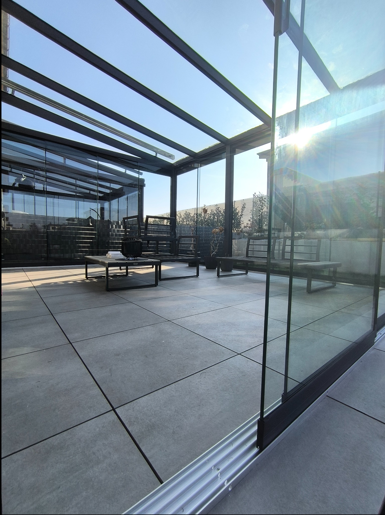 Szklana zabudowa tarasu z metalową konstrukcją nośną, widoczny system przesuwny szyb, meble ogrodowe i dekoracje na stoliku kawowym.