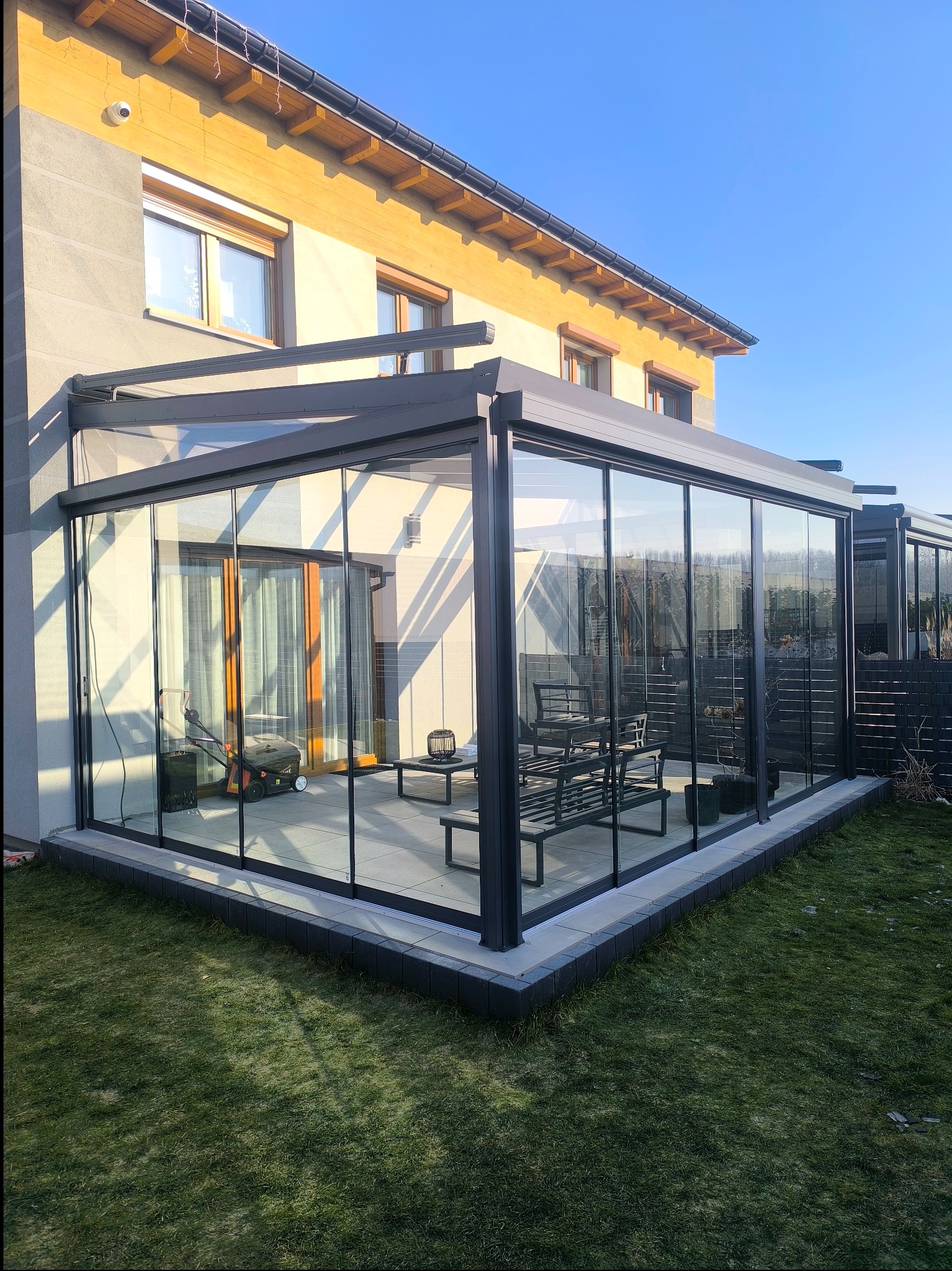 Szklana zabudowa tarasu z przesuwnymi drzwiami, widoczne meble ogrodowe i kosiarka w środku, na zewnątrz trawnik i fragment domu w słoneczny dzień.