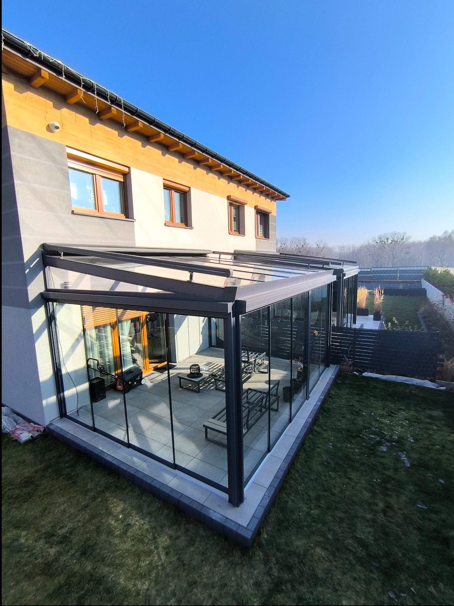 Nowoczesna, przeszklona konstrukcja tarasowa z metalową ramą przylegająca do domu, widok z zewnątrz na ogród i błękitne niebo.