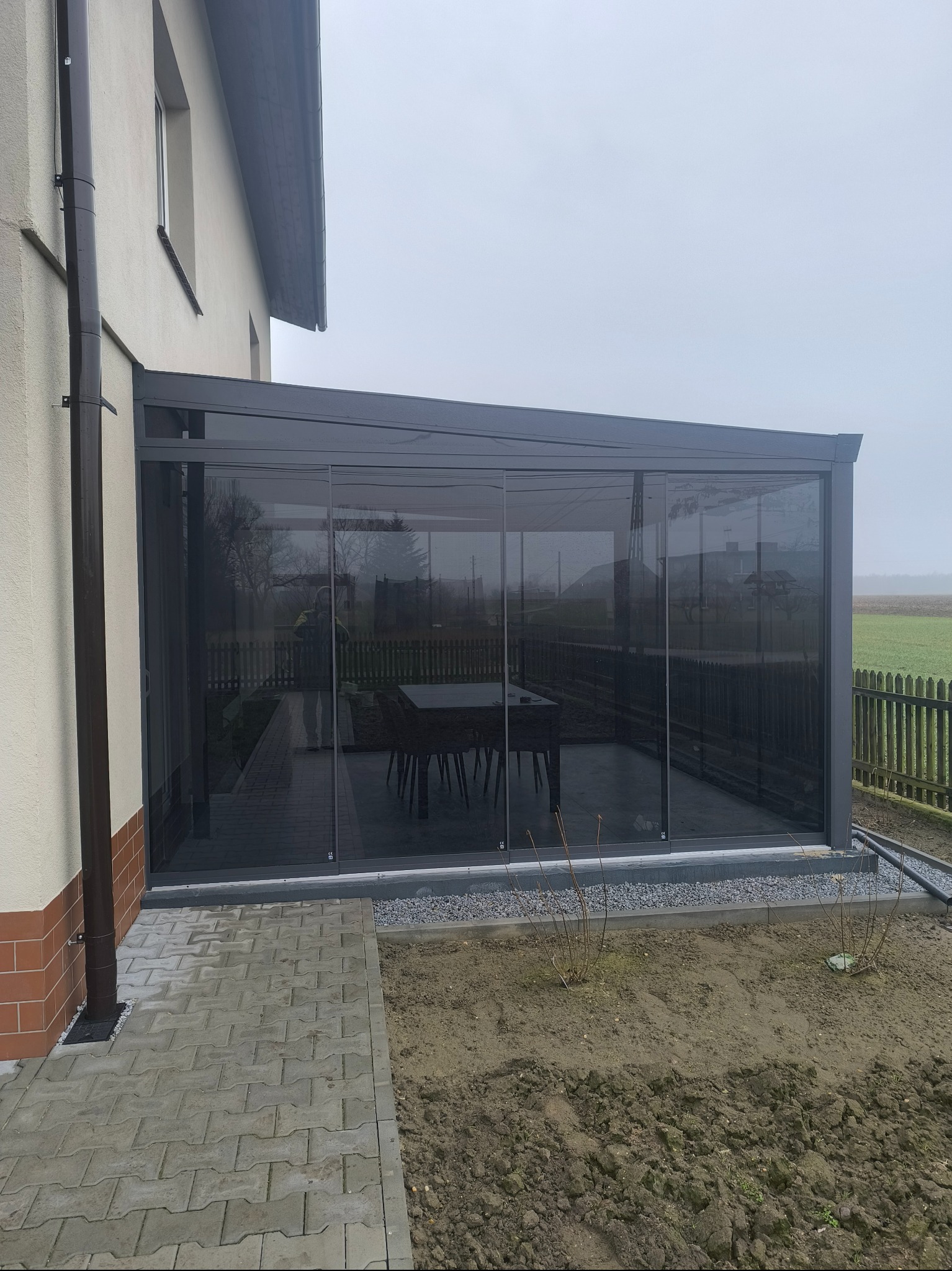 Szara, przeszklona konstrukcja przylegająca do budynku z widocznym stołem i krzesłami wewnątrz, obok kostka brukowa i rabata z ziemią.