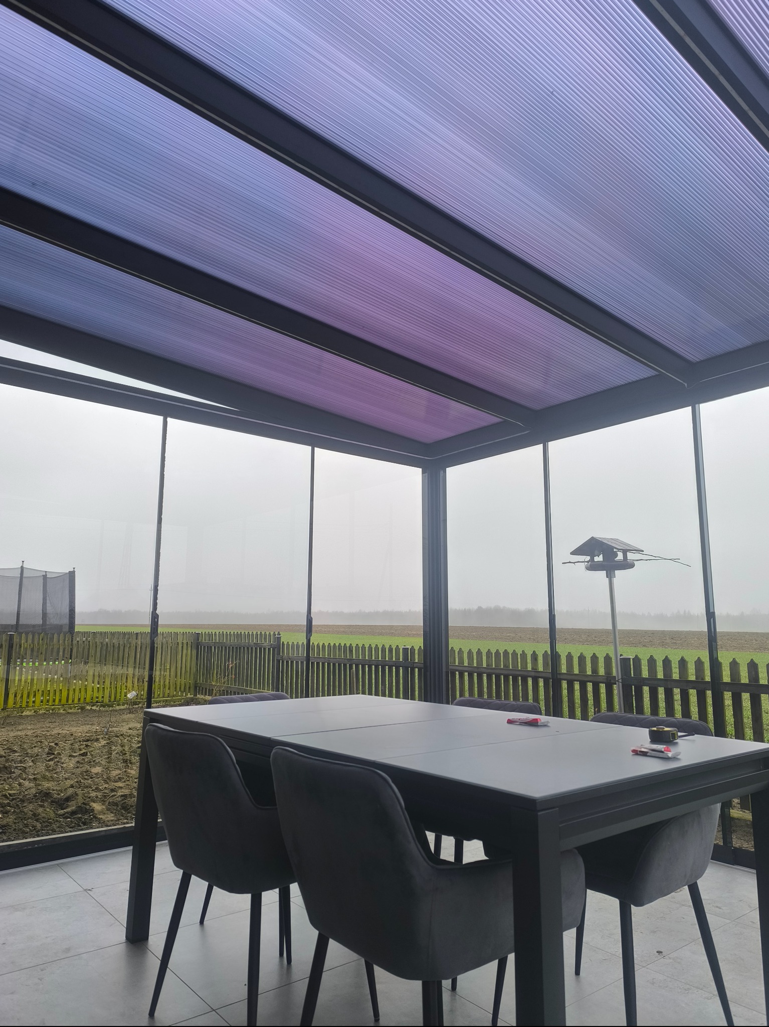 Zadaszony taras z metalową konstrukcją i przezroczystym dachem, widok na ogród z drewnianym płotem i trampoliną w tle.