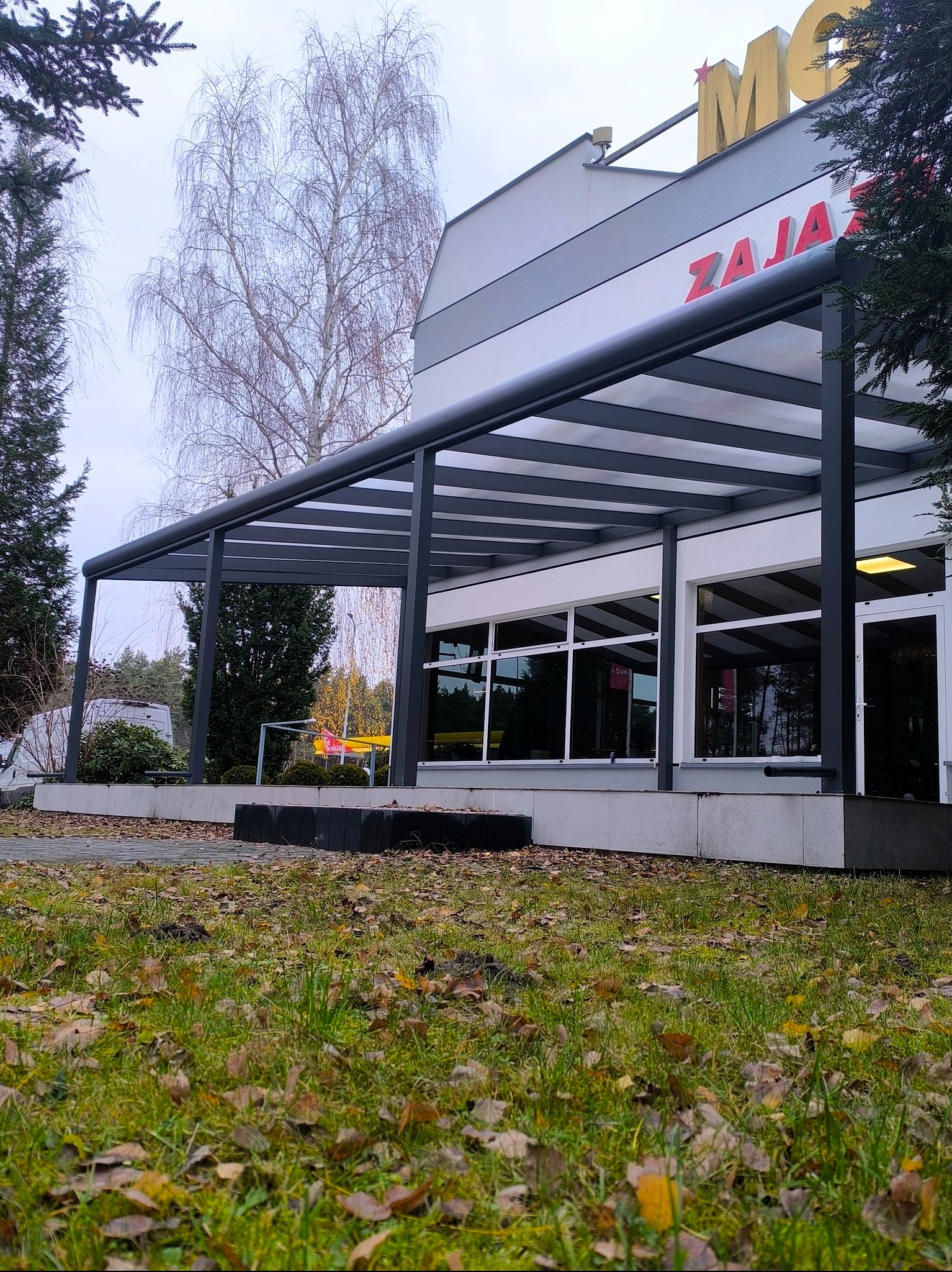 Metalowa pergola w kolorze antracytowym przylegająca do budynku z dużymi oknami, widok z poziomu trawnika pokrytego opadłymi liśćmi.