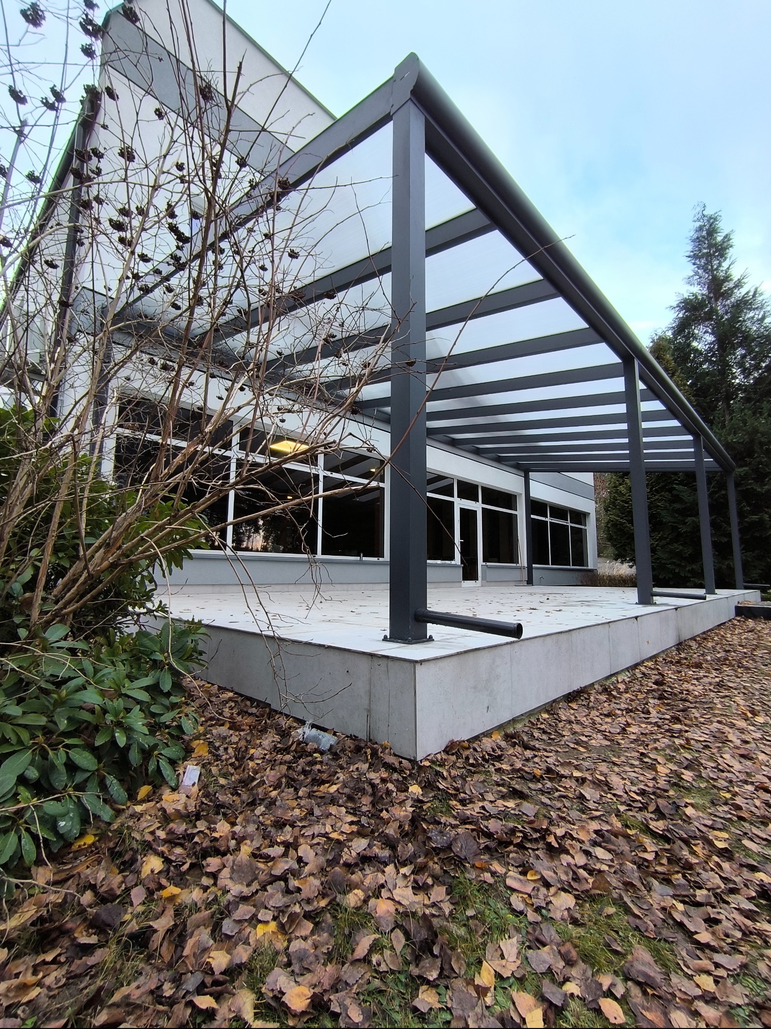 Nowoczesna, szara konstrukcja z metalu i przezroczystego zadaszenia, prawdopodobnie pergola, przylegająca do budynku z dużymi oknami, na tle jesiennego krajobrazu z opadłymi liśćmi.