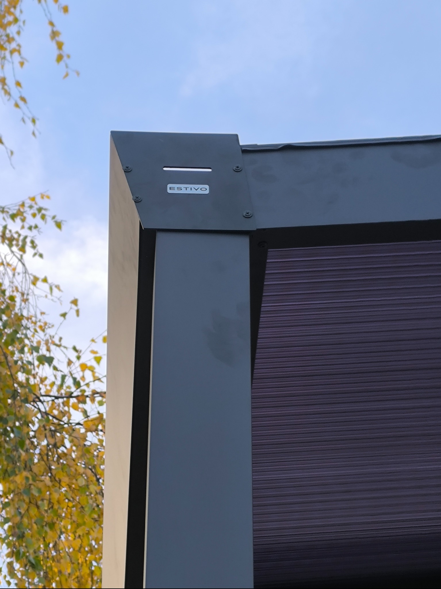 Fragment narożnika metalowej pergoli z widocznym logo 'ESTIVO', na tle błękitnego nieba i drzewa z żółtymi liśćmi.