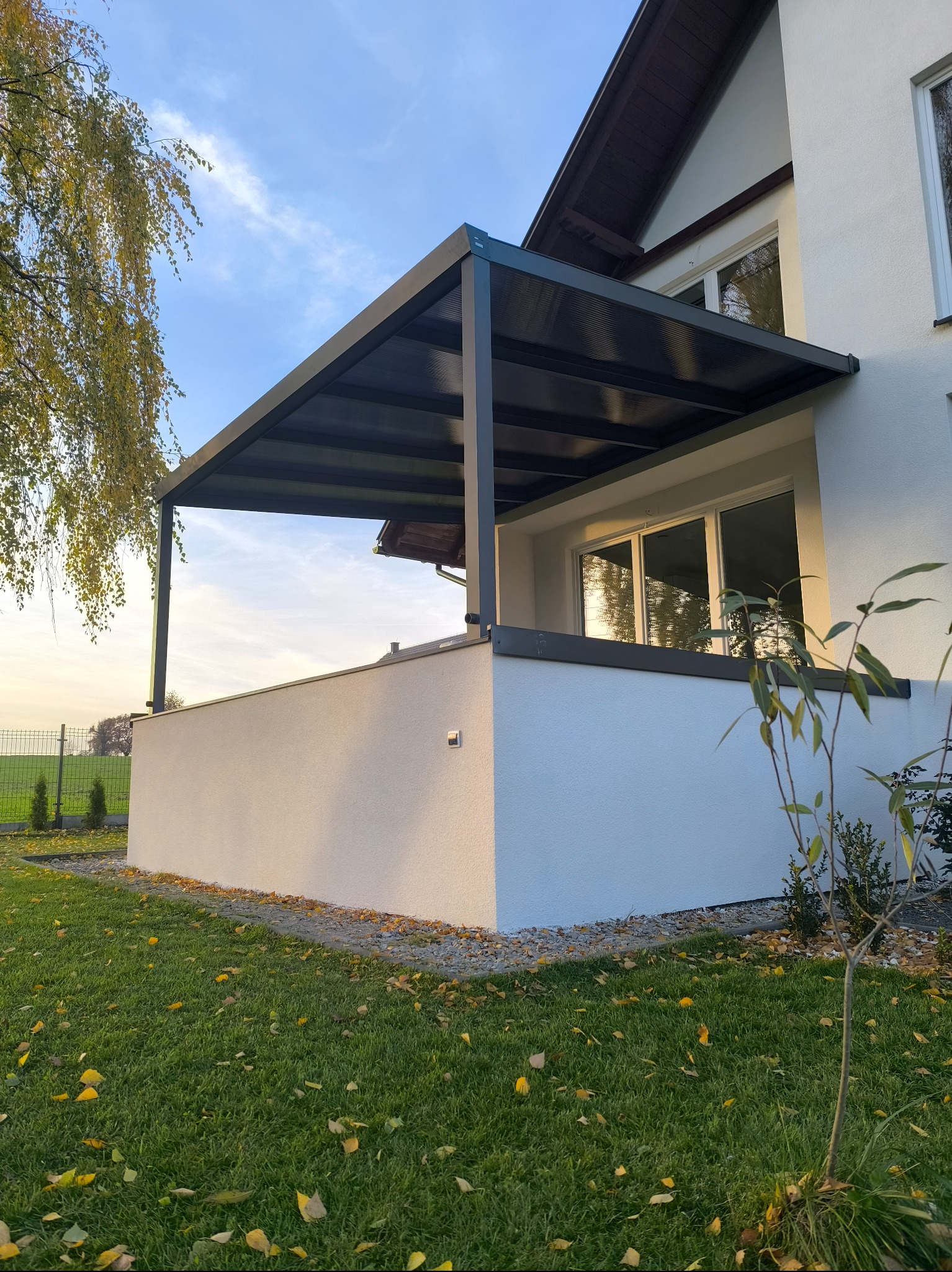 Nowoczesna, grafitowa pergola przy białym domu jednorodzinnym, widok z trawnika z opadłymi liśćmi, błękitne niebo w tle.